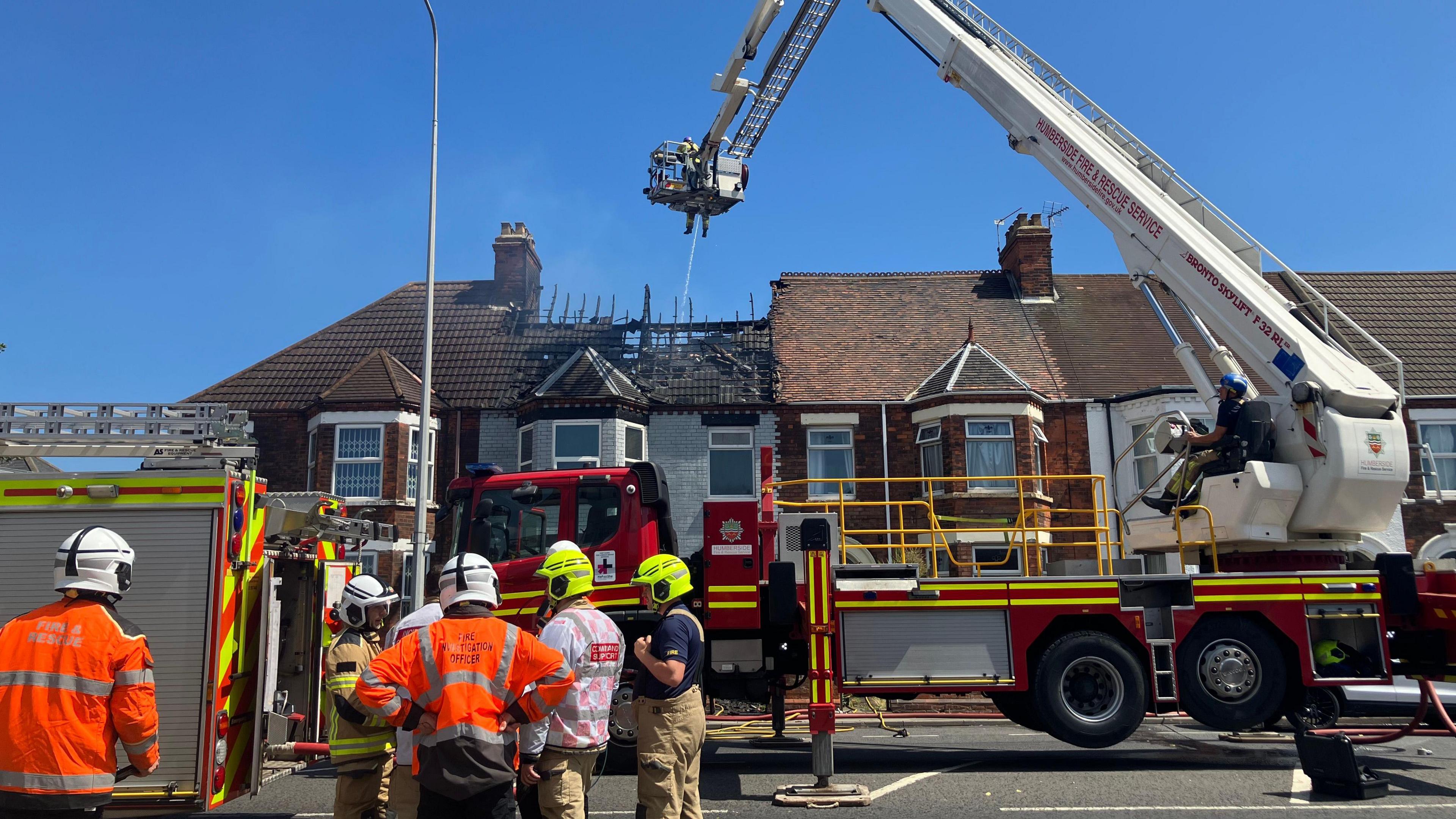 Five fightfighters in uniform with an aerial platform with a firefighter on it hosing down the fire, which is inside a terraced property with the roof burnt off.
There is a second fire engine in shot. The house is amongst a terraced row of houses. The sky is clear.