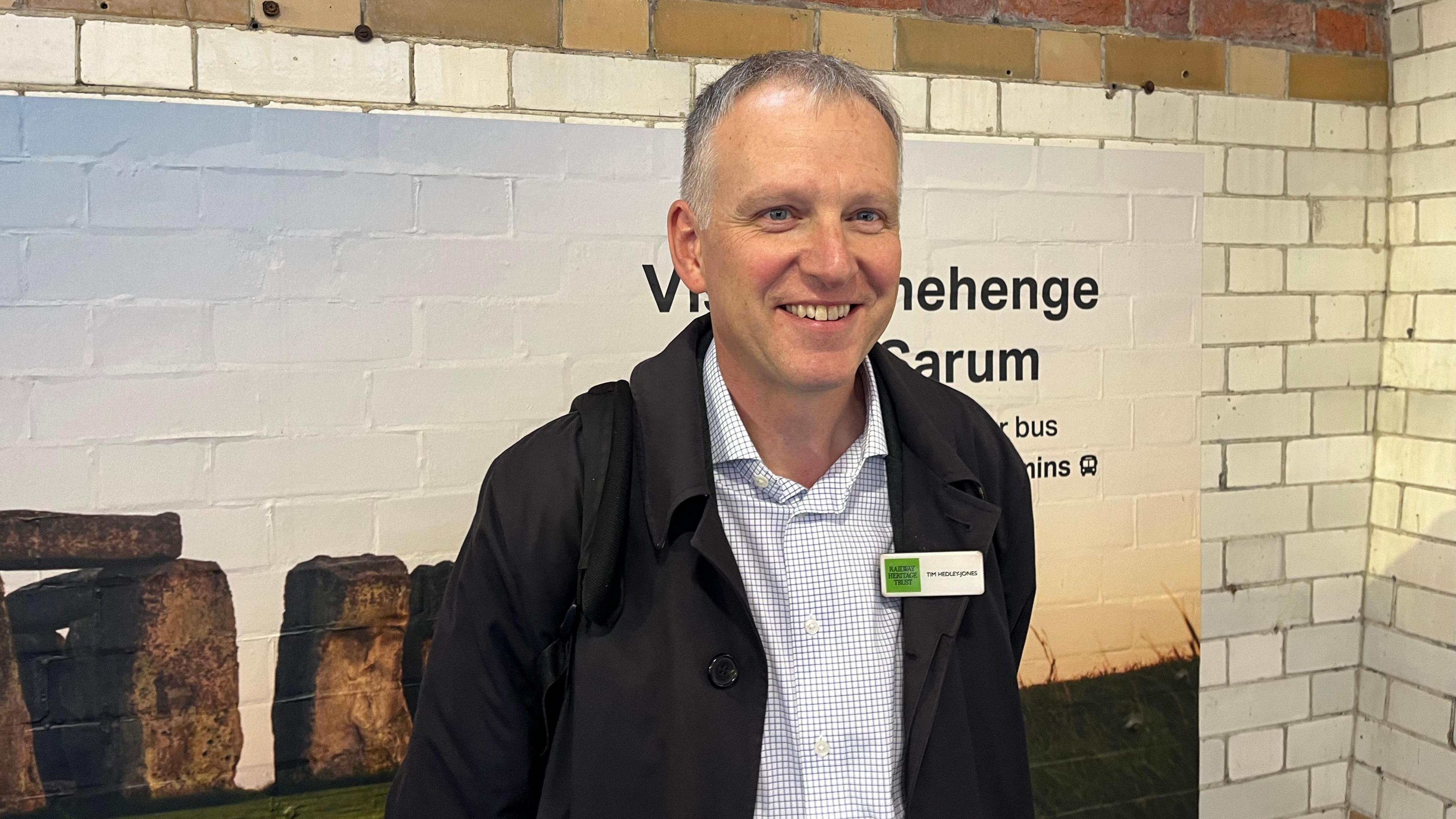 Tim Hedley-Jones smiles while standing in front of subway tiles and a big picture of Stonehenge pointing to where the tour buses are