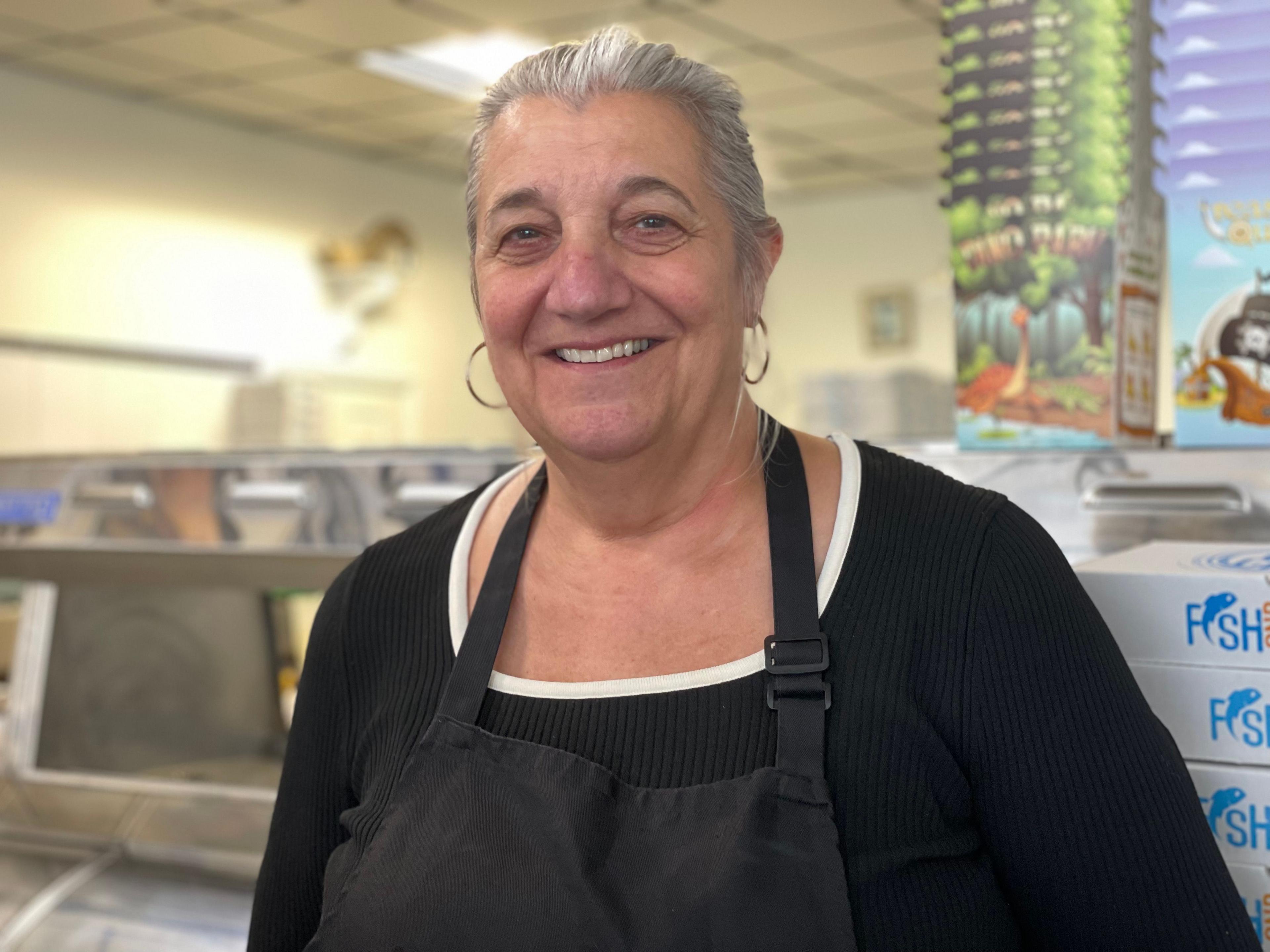 Patricia Paiva De Brito wearing a black top and apron stood in front of the range in her chip shop