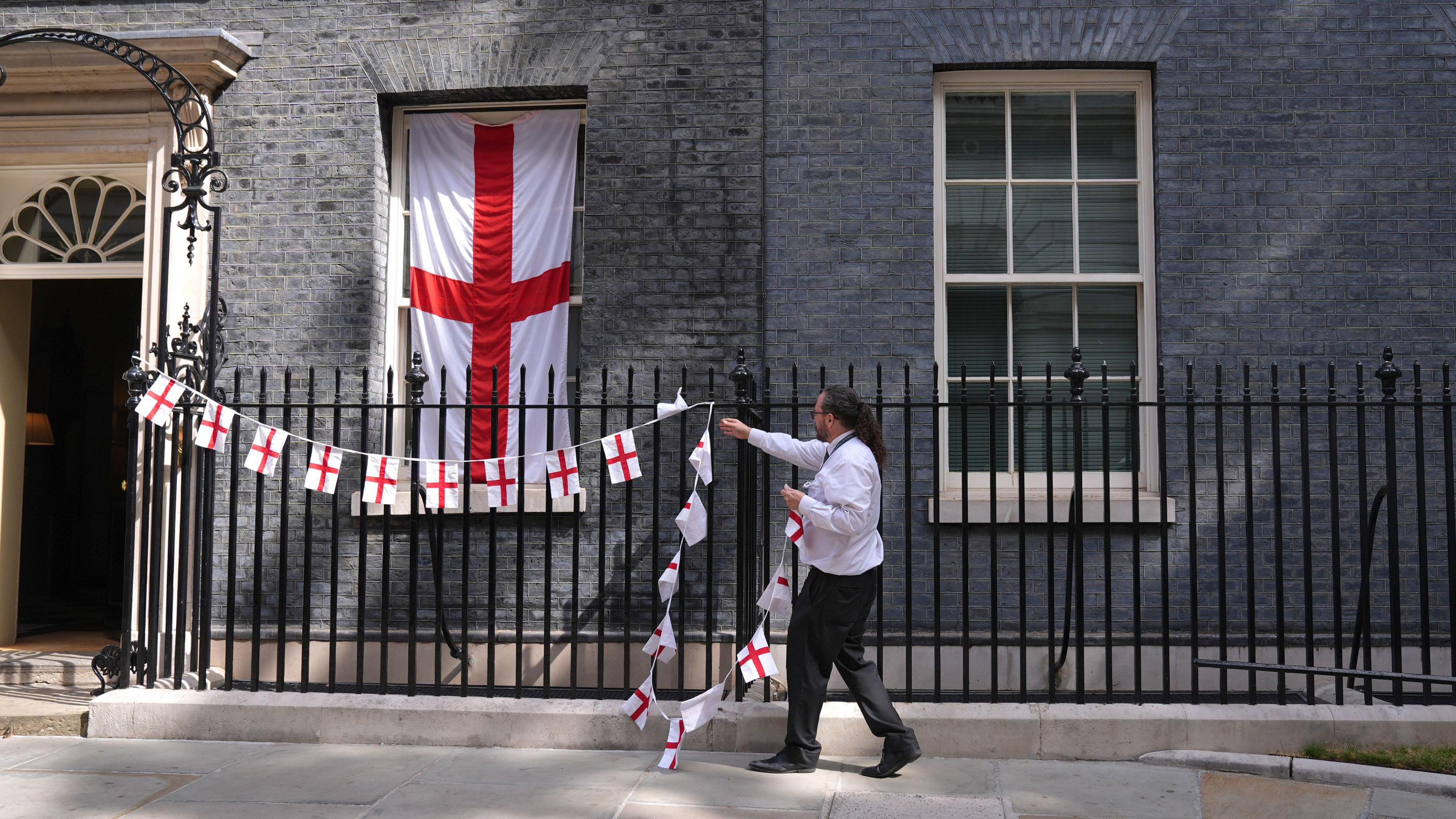 A man in a shirt and black trousers takes down England flag bunting that was on the railings outside No 10 Downing Street. A large England flag still remains draped in front one of the windows.