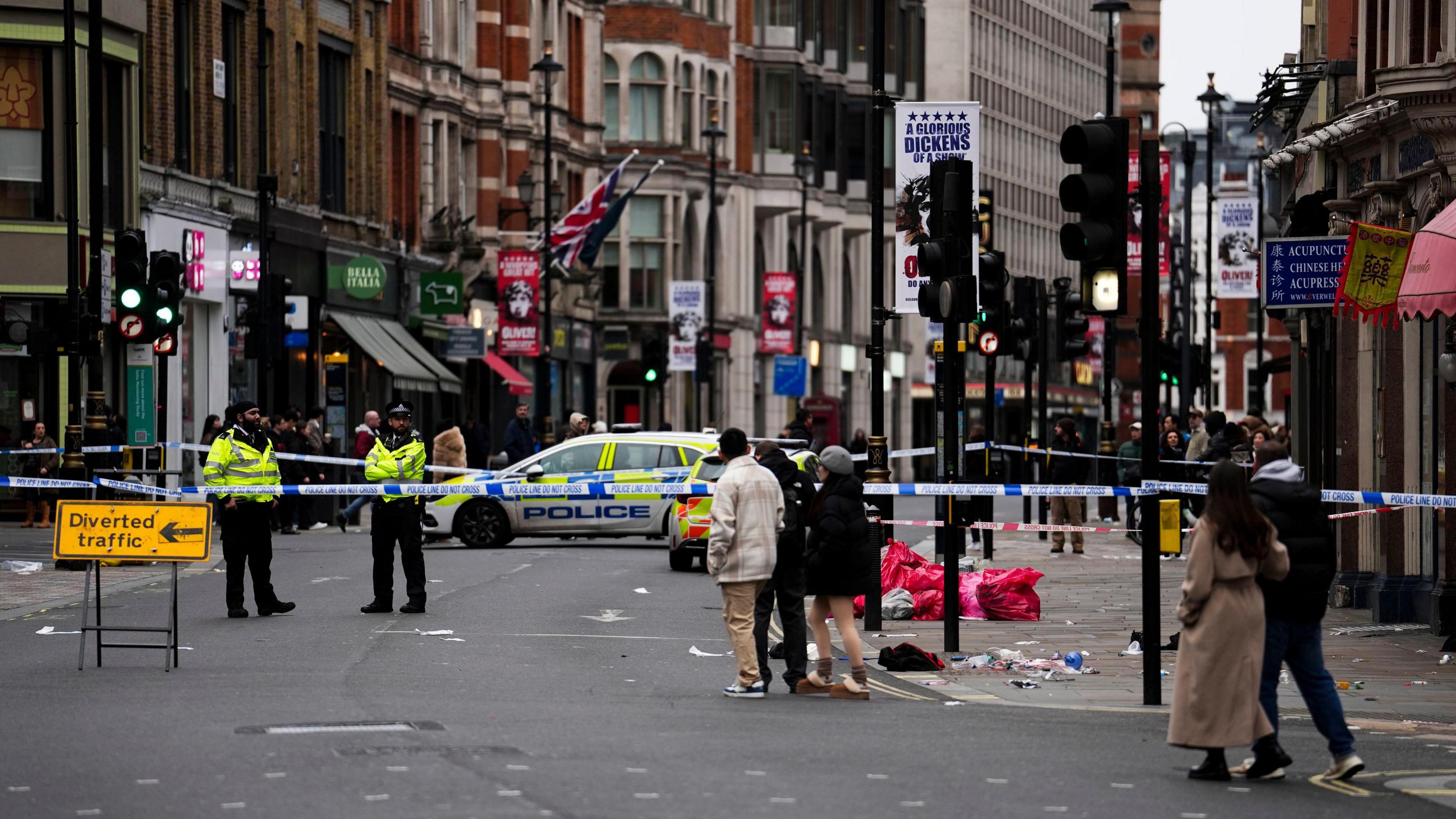 Members of the public walk past a police cordon on Shaftesbury Avenue. Two police officers are standing behind the cordon. There is a yellow sign that says 'diverted traffic' and an arrow pointing left which has been placed in front of the cordon.