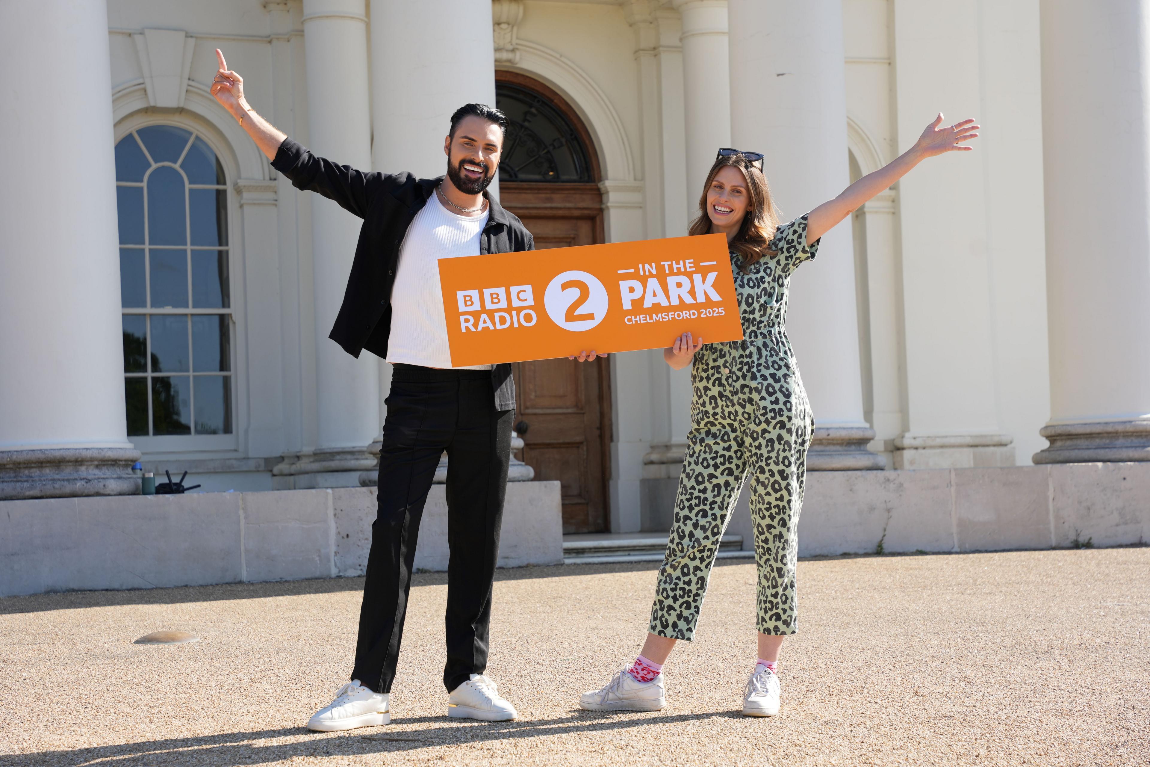 Rylan Clark and Ellie Taylor stand in front of Hylands House, holding an orange sign with white writing which reads "BBC Radio 2 in the Park Chelmsford 2025". Rylan is stood on the left of frame, wearing black trousers and a black open shirt with white t-shirt underneath. He has dark hair and a beard. Ellie stands on the right of frame, wearing a leopard-print boiler suit, and has dark sunglasses pushed on top of her head. She has brown hair below shoulder-length. Both Rylan and Ellie are holding the sign with one hand and their other arm is spread wide. Hylands House behind them is a large white building with white columns.