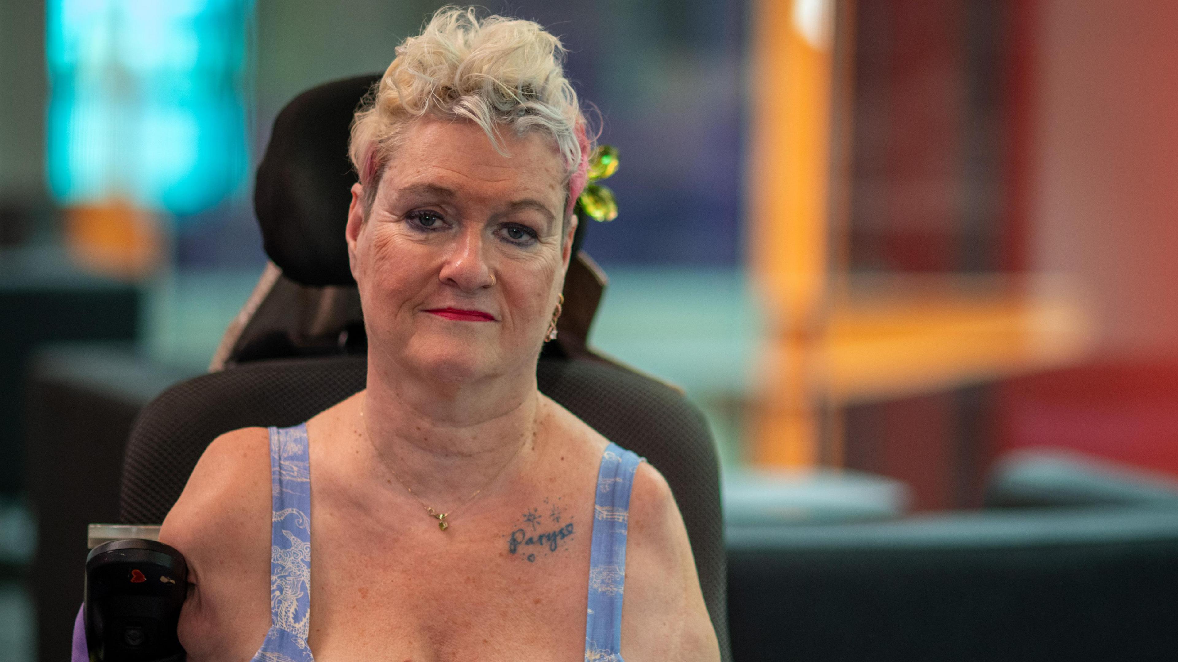 Alison Lapper is staring directly at the camera. She is wearing a blue outfit and has a pink and green hair clip in her blonde hair. Her son's name is tattooed above her heart.  