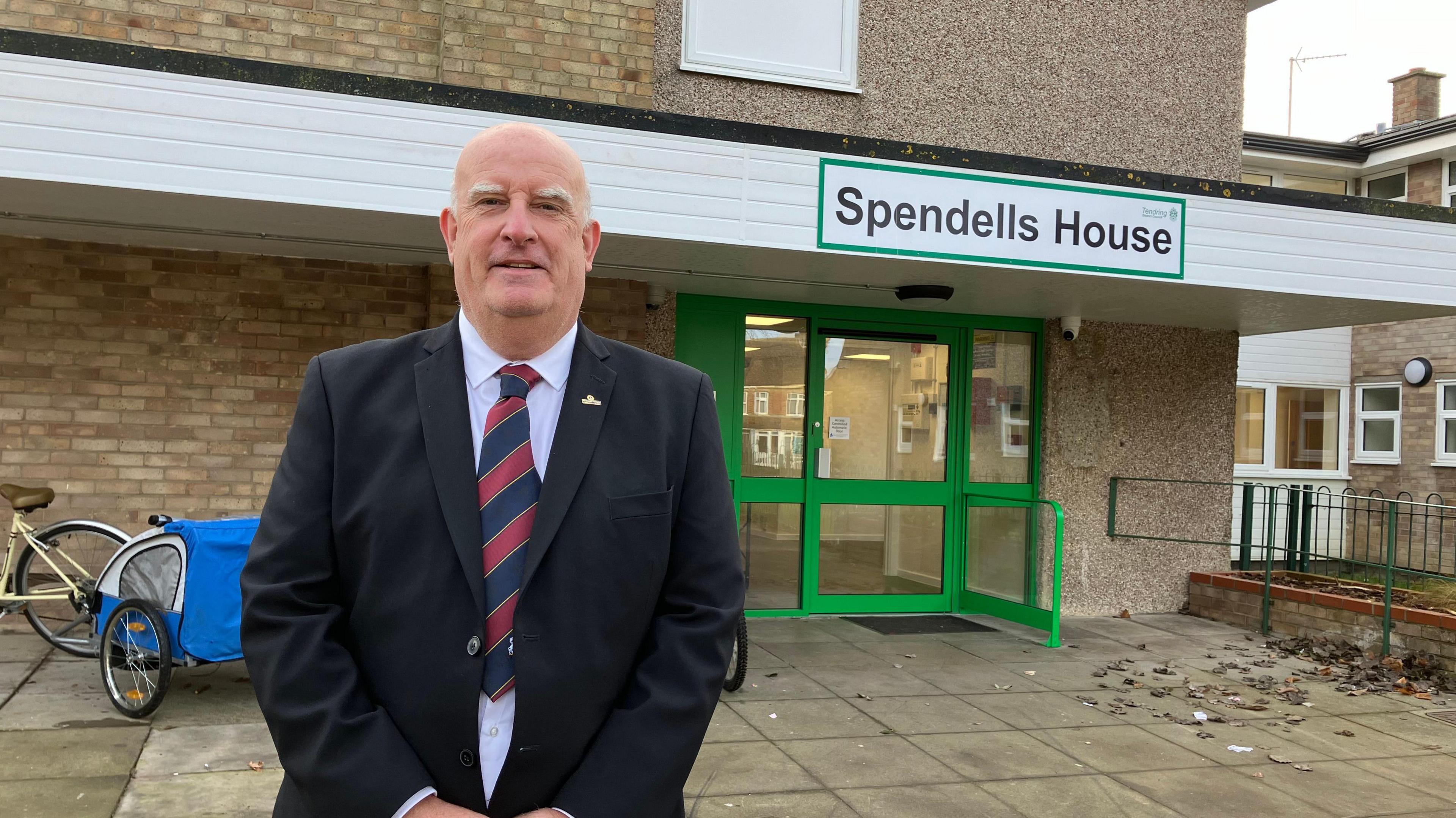 A man in a three-piece suit smiling in front of a building called "Spendells House"