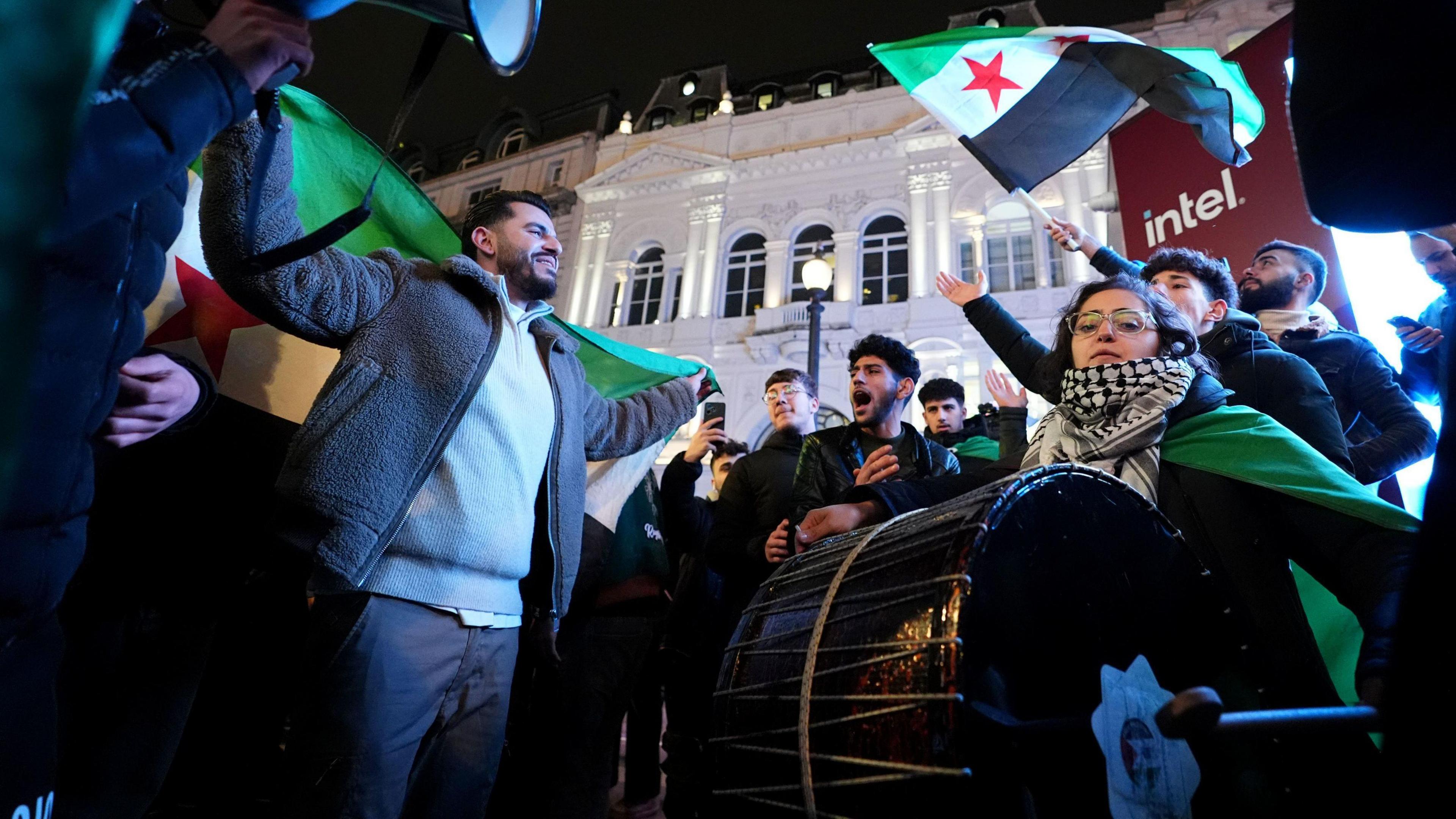 A man holding a Syrian flag and a woman playing traditional Middle Eastern drums.