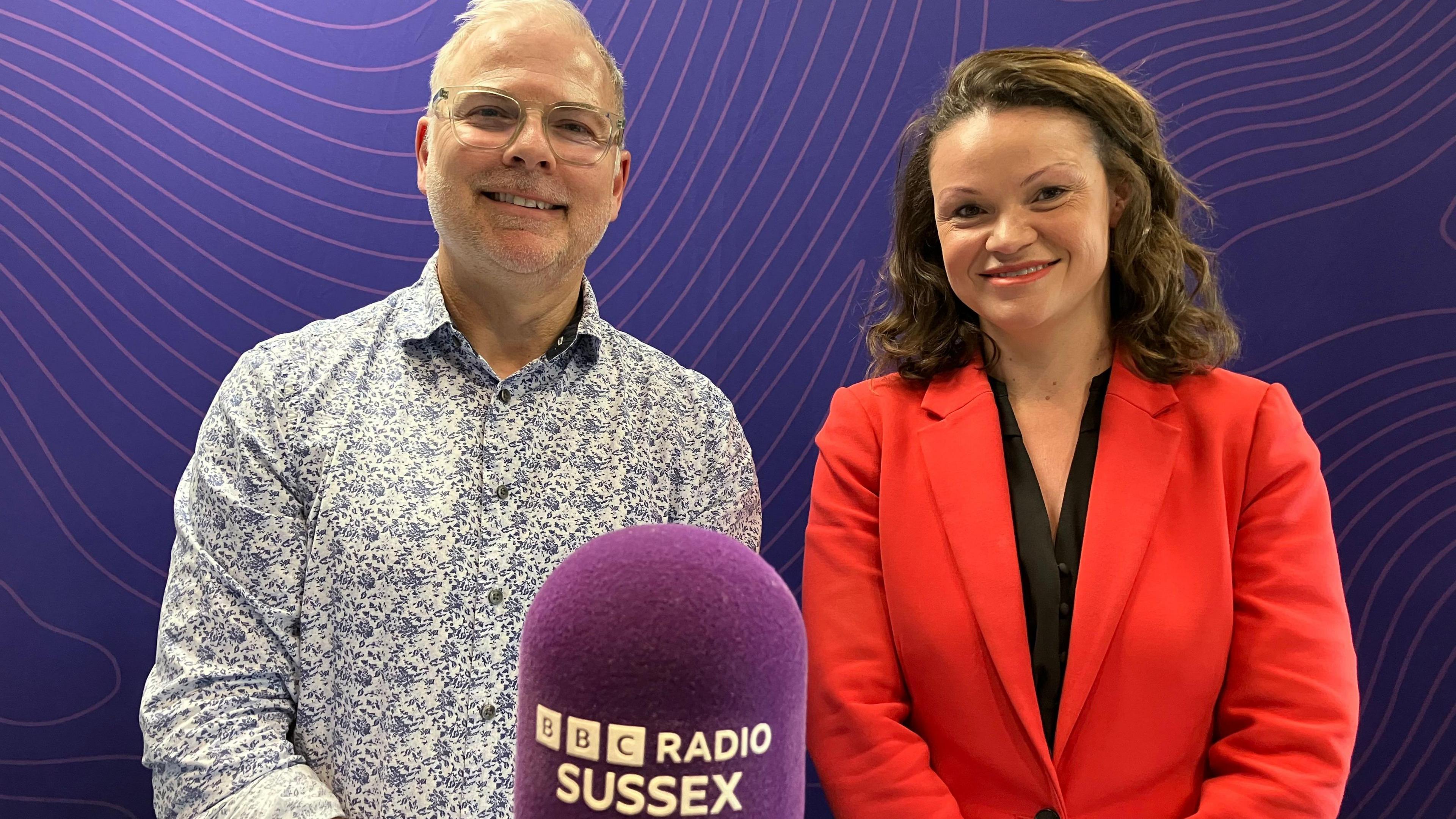 On the left presenter Danny Pike has grey hair and a grey stubble beard. He wears glasses and a patterned white and blue shirt. On the right council leader Bella Sankey has long brown wavy hair and wears a bright red suit jacket over a black top. This photo is taken in a radio studio with a purple background and a purple microphone in the foreground with the BBC Radio Sussex logo on it in white writing.