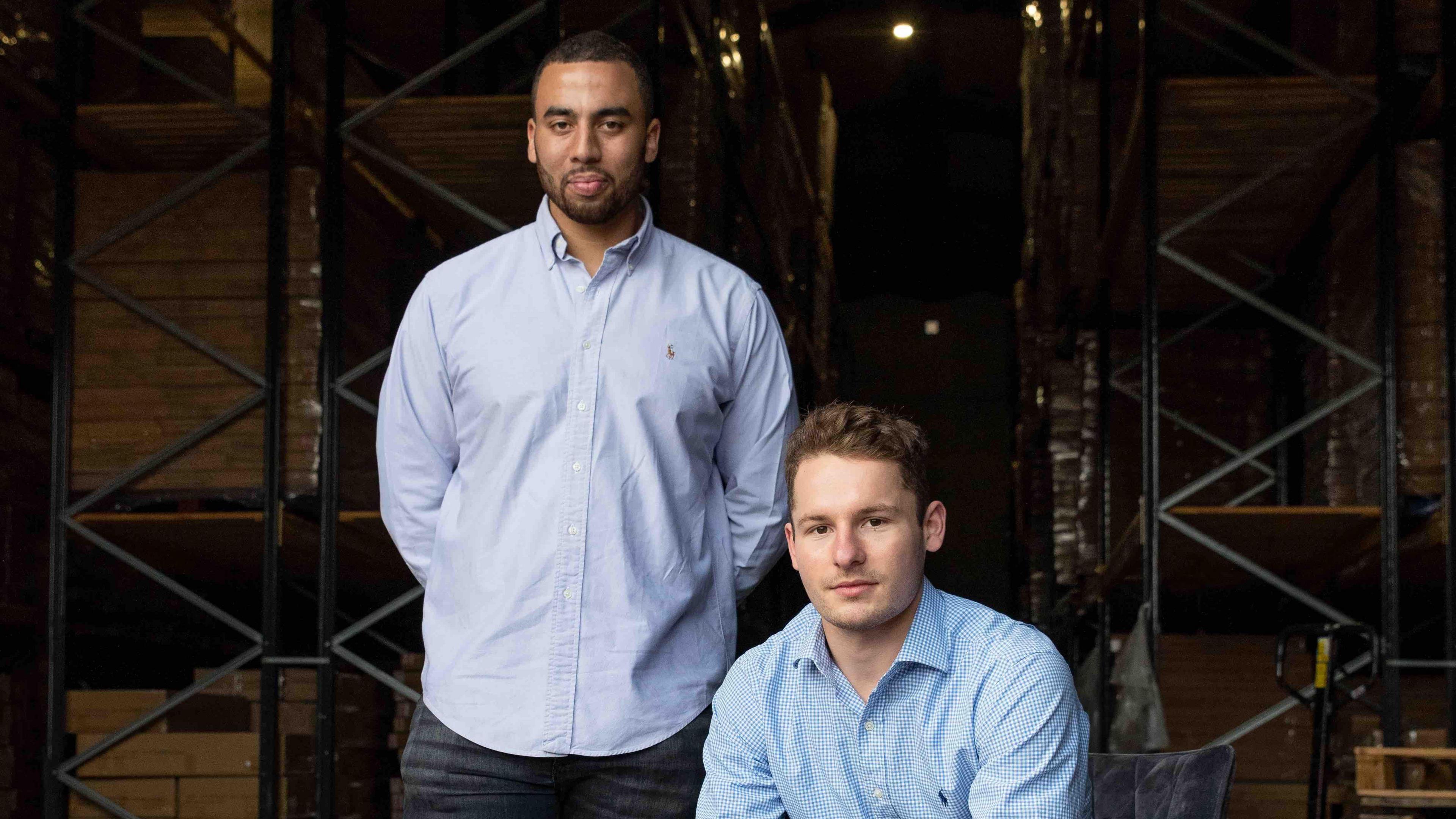 Dan Beckles and Money George together in a warehouse, looking at the camera. Both wear smart blue shirts, Dan stands and Monty is sitting.