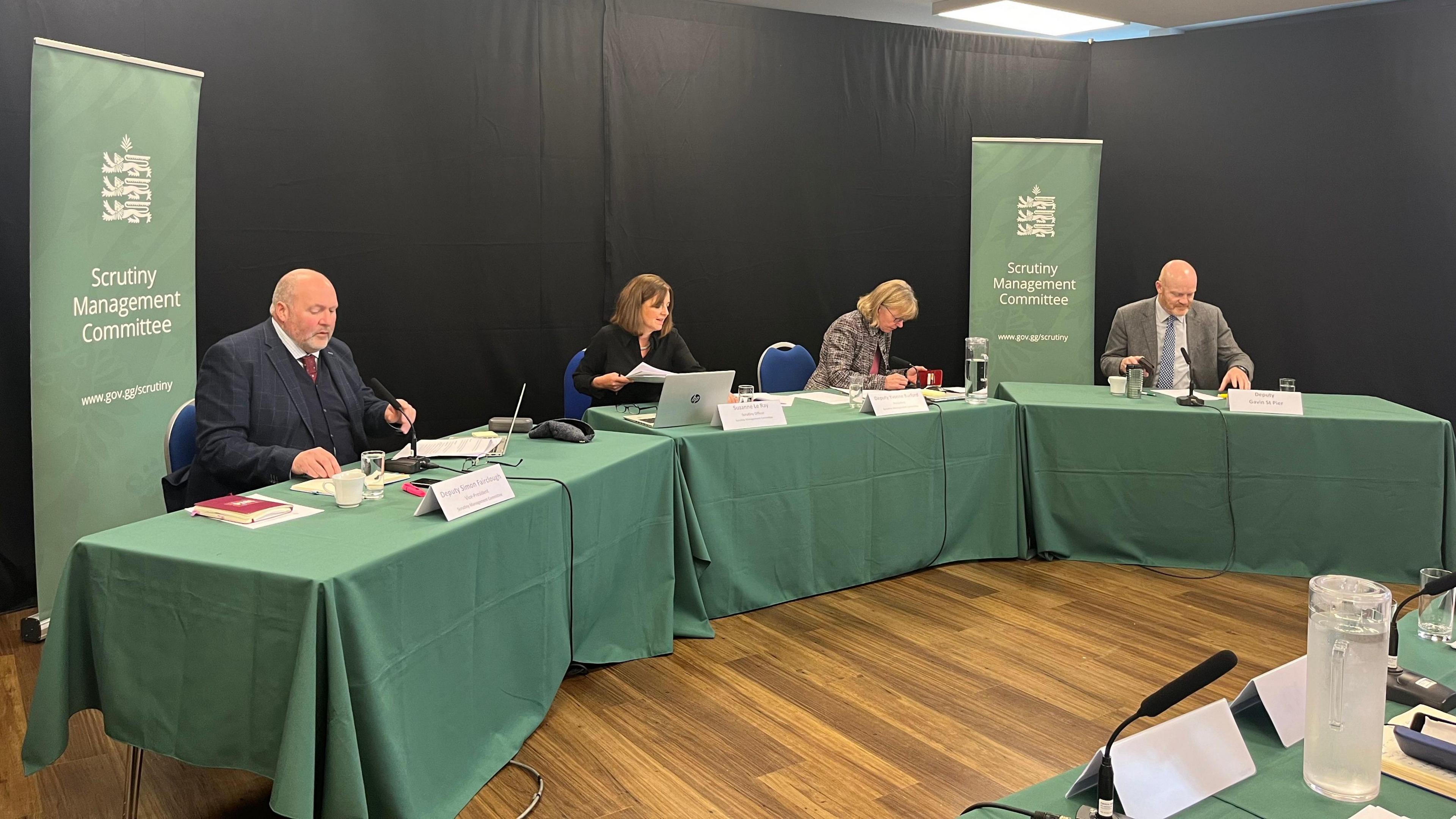 Four people sitting at a table. Two signs saying Scrutiny Management Committee