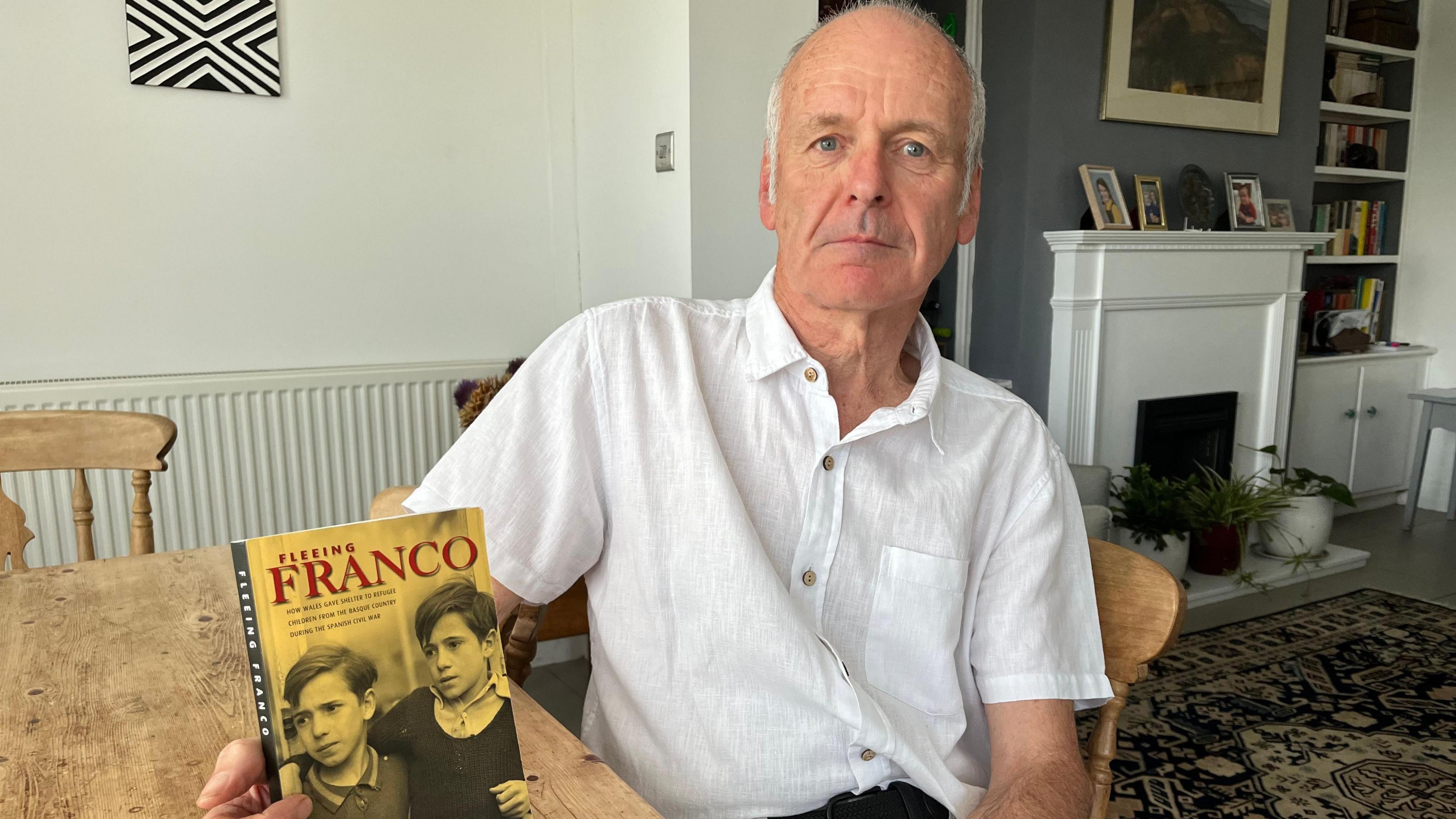 An older man looks at the camera. He is bald but has white hair on the sides. He is wearing a white shirt and is sitting at a table holding a book which has the image of two boys on it and the title "Fleeing Franco". Behind is his living room, where there is a fire place and various pictures framed.