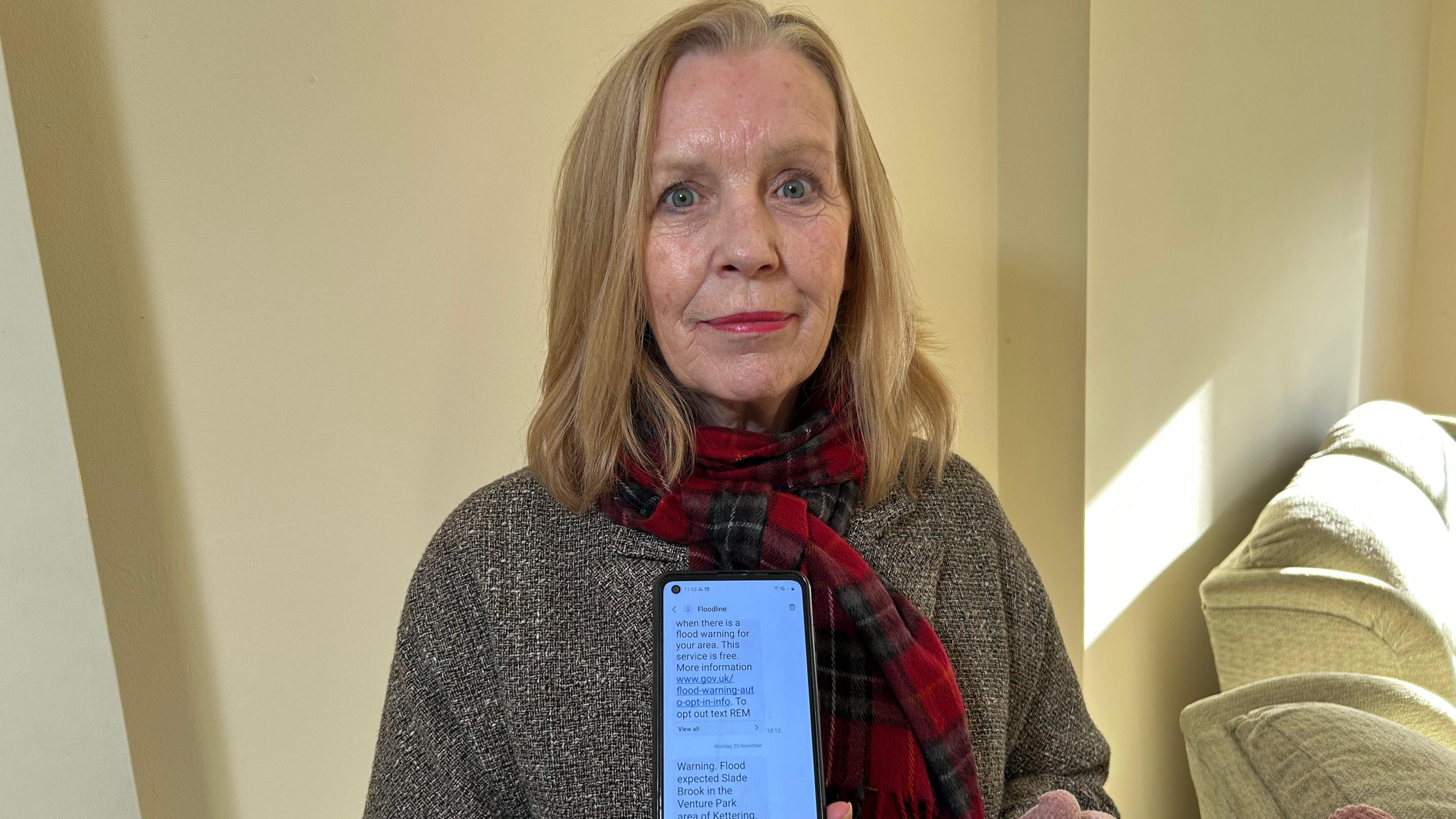 A woman with shoulder length blonde hair smiles at the camera while showing her phone with a flood alert on it. She is standing in a living room while wearing a grey coat and tartan scarf.