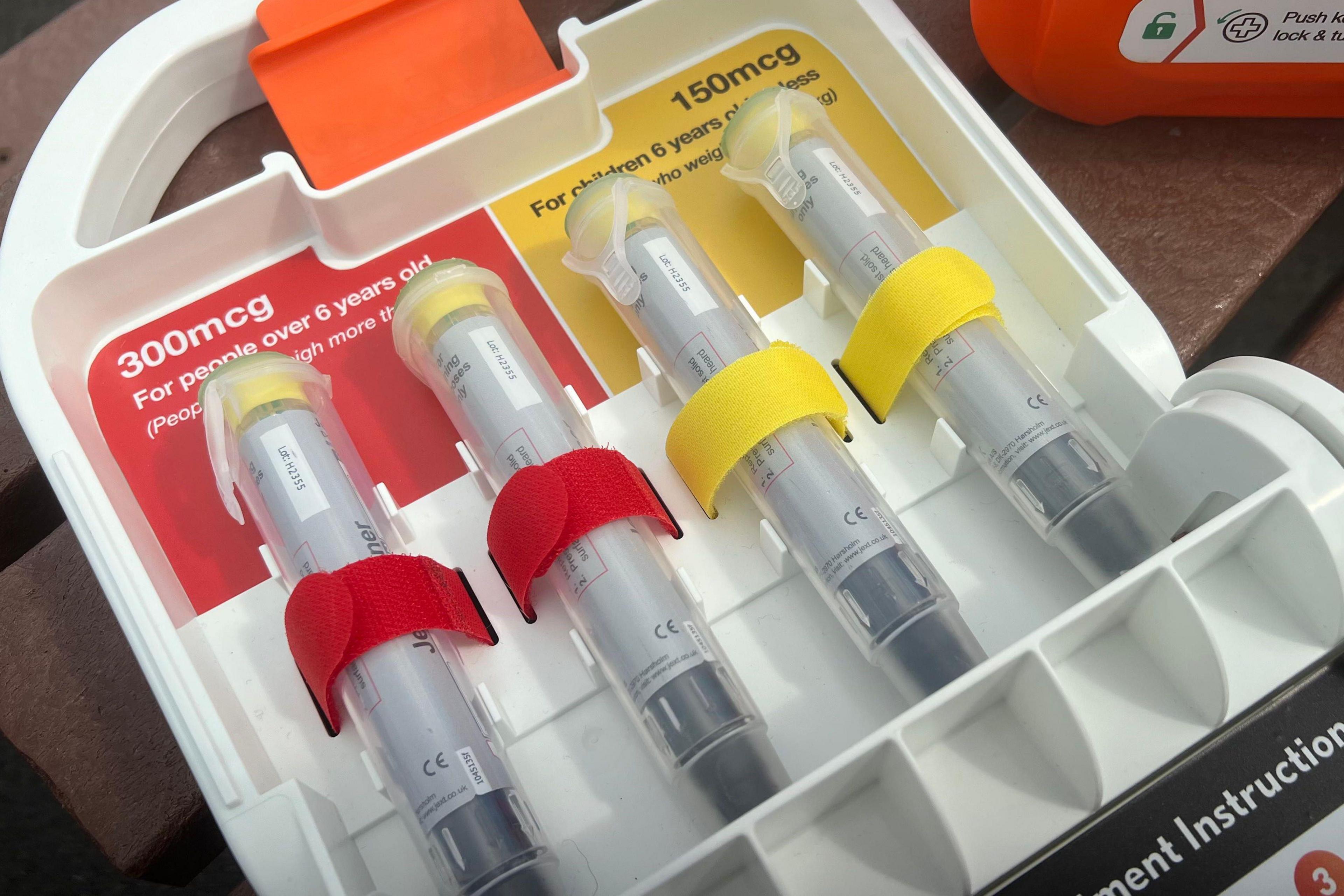 A white wall-mounted emergency allergy kit containing four adrenaline pens. Two are secured with red Velcro labelled “300 MCG” for people over six years old, and two with yellow Velcro labelled “150 MCG” for those under six.