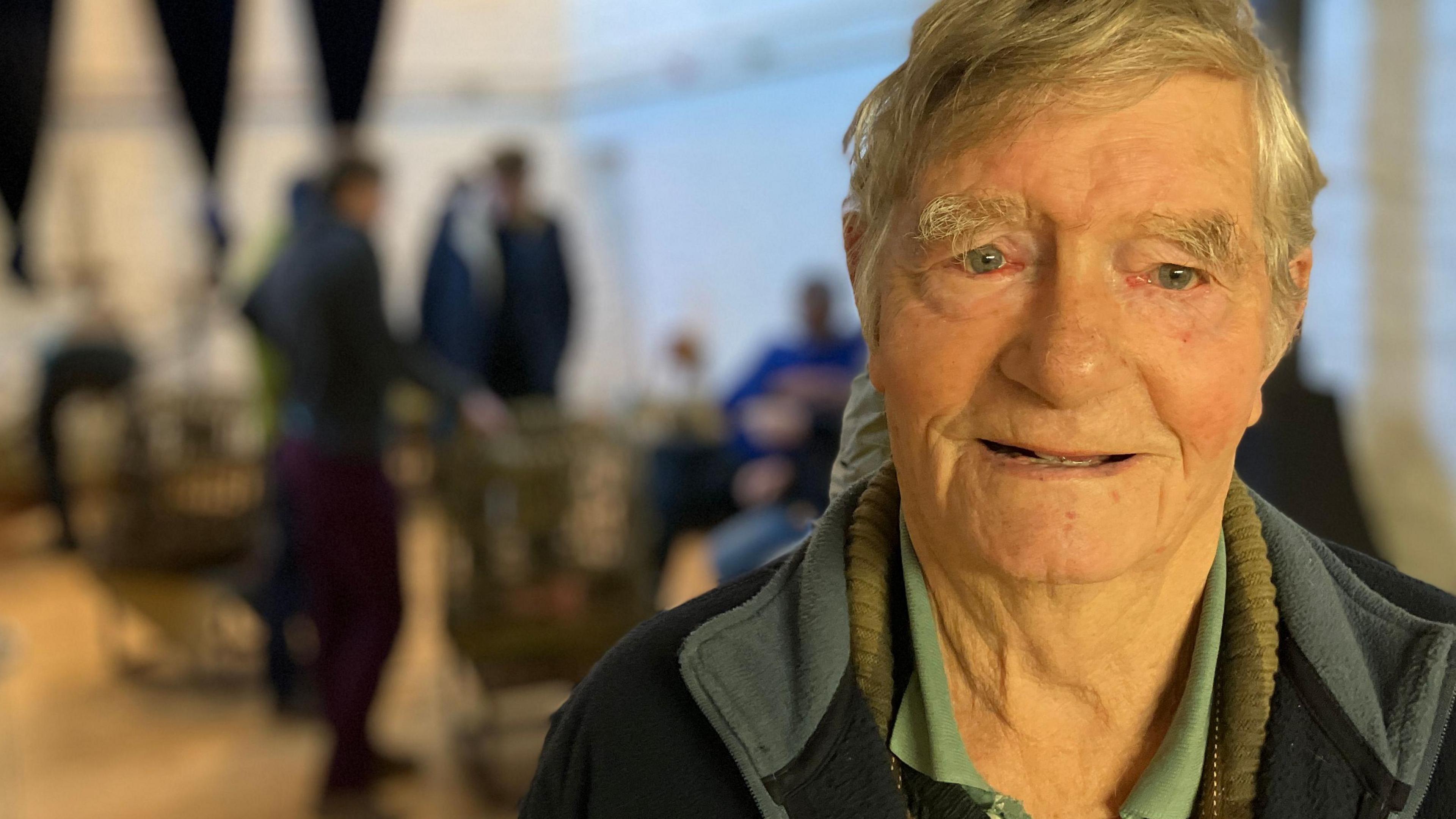 An elderly man with fair hair wearing a dark top and green shirt.