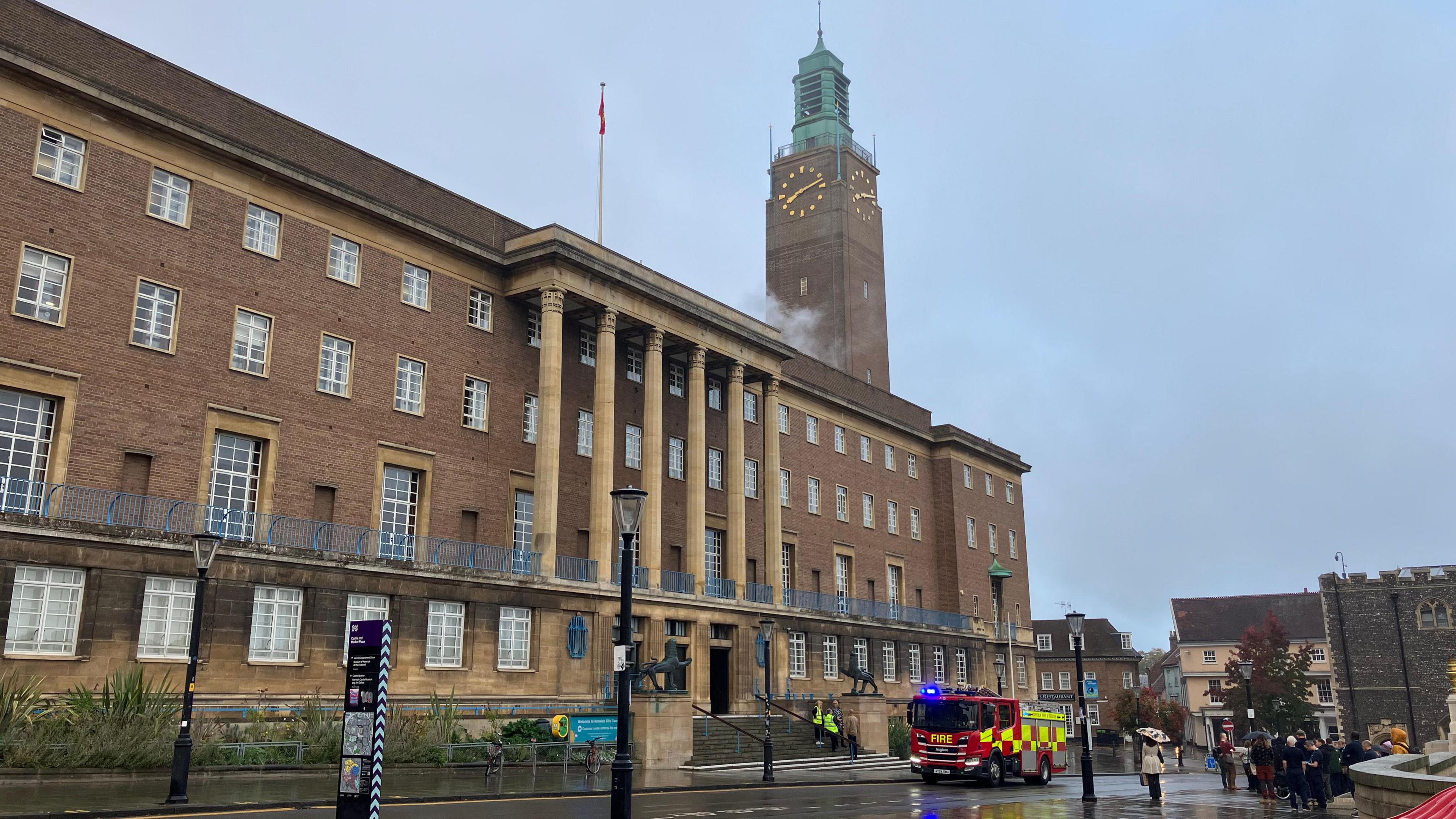 Wisps of smoke can be seen near a clock tower on the roof of City Hall in Norwich. It is an imposing civic building that is four storeys high. It has six columns above a flight of steps leading to the entrance. A fire engine is parked on the rain-soaked street outside.