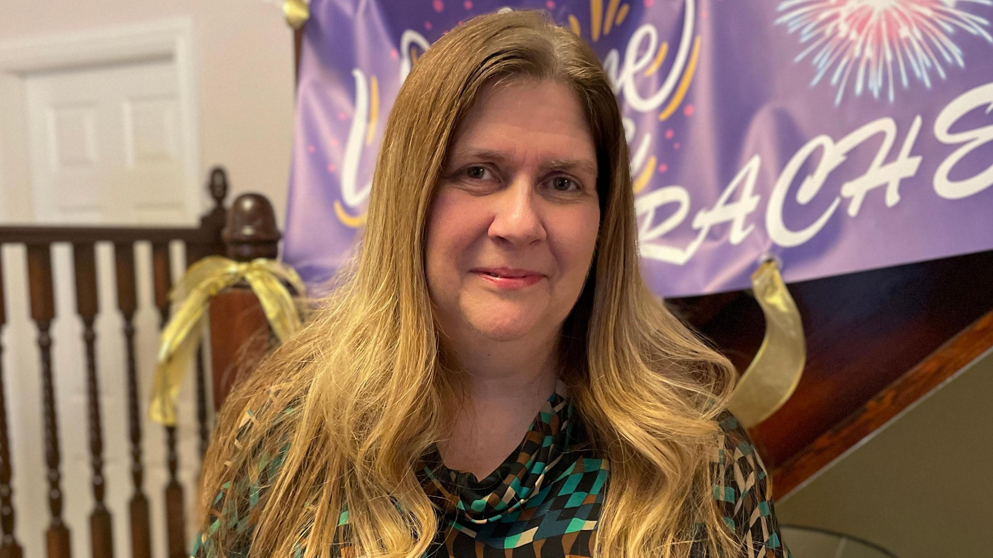 A woman with shoulder length fair hair and a green patterned top stands in front of a purple banner