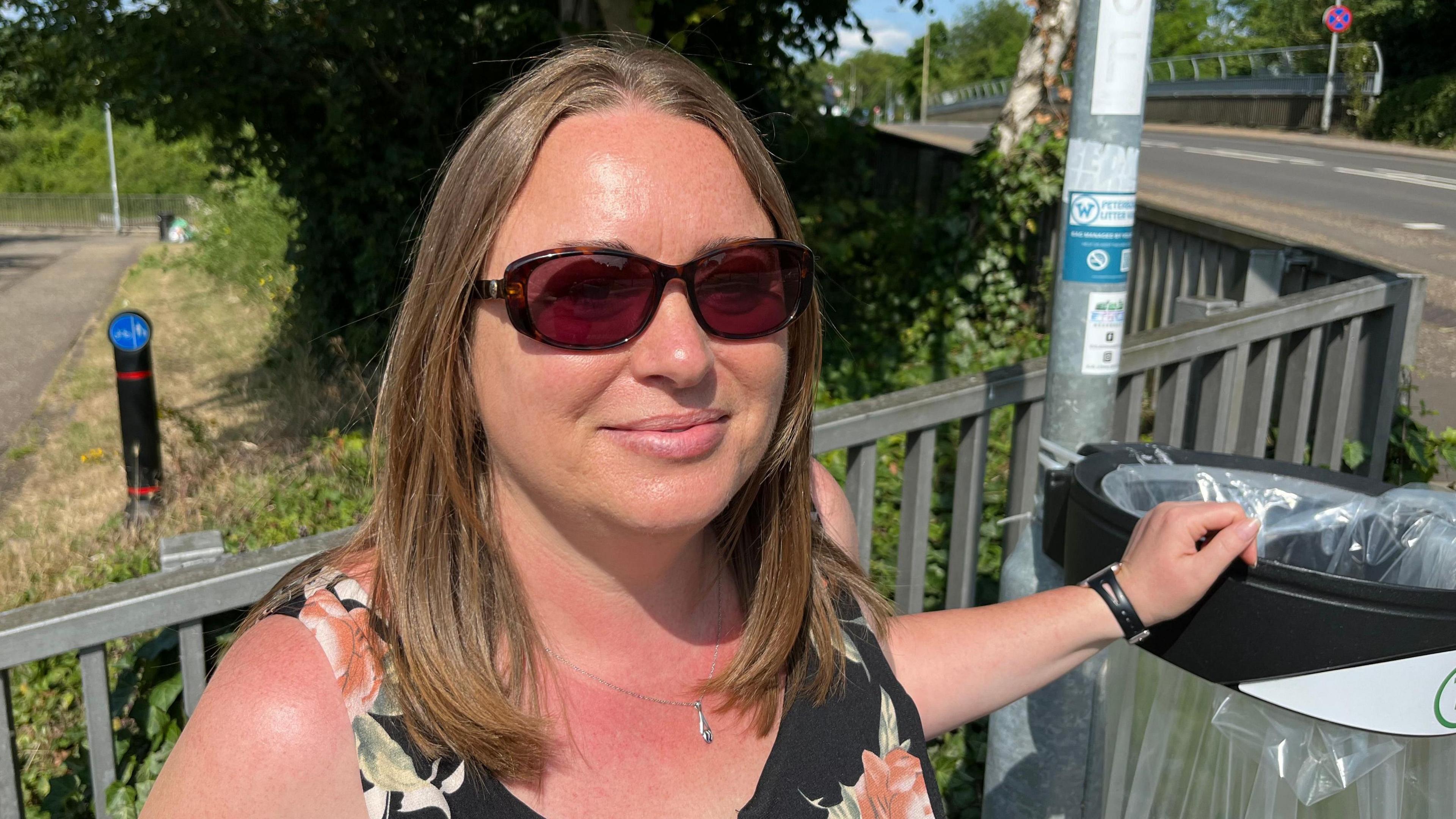 Samantha Dalton, 48, has brown/light blonde shoulder length hair parted in the middle of her head. She has dark glasses and a silver chain around her neck. She is wearing a dark floral sleeveless dress and has her hand on the rim of a public bin. Behind her are railings and roadways.