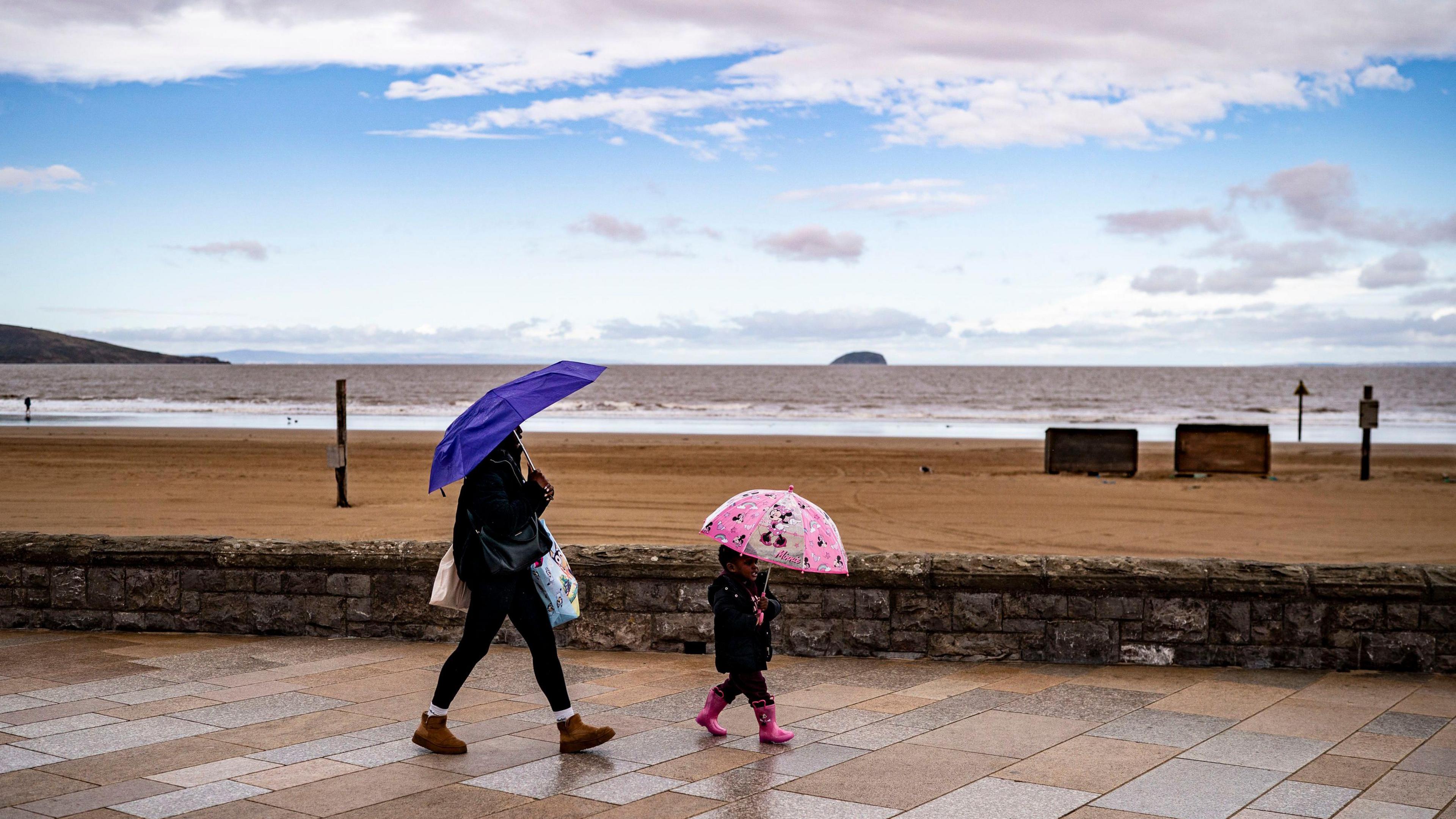 A woman and a child walk along a seafront holding umbrellas, with the beach and the sea in the background and a sky of blue and grey overhead
