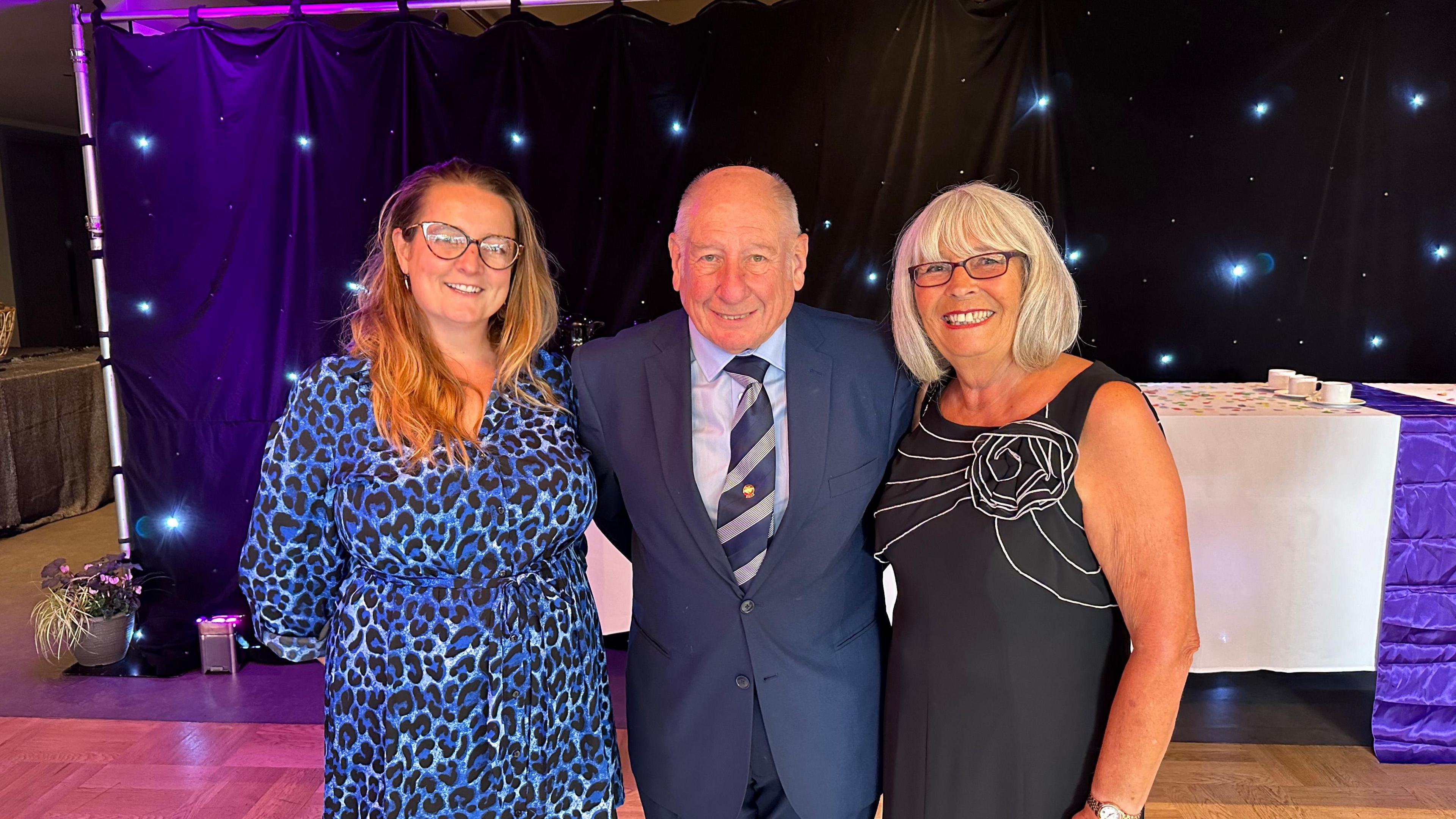 Arthur Harman is in the middle between two ladies smiling as he wins an award for his volunteering. He is wearing a dark blue suit with a grey and blue tie with a blue shirt.