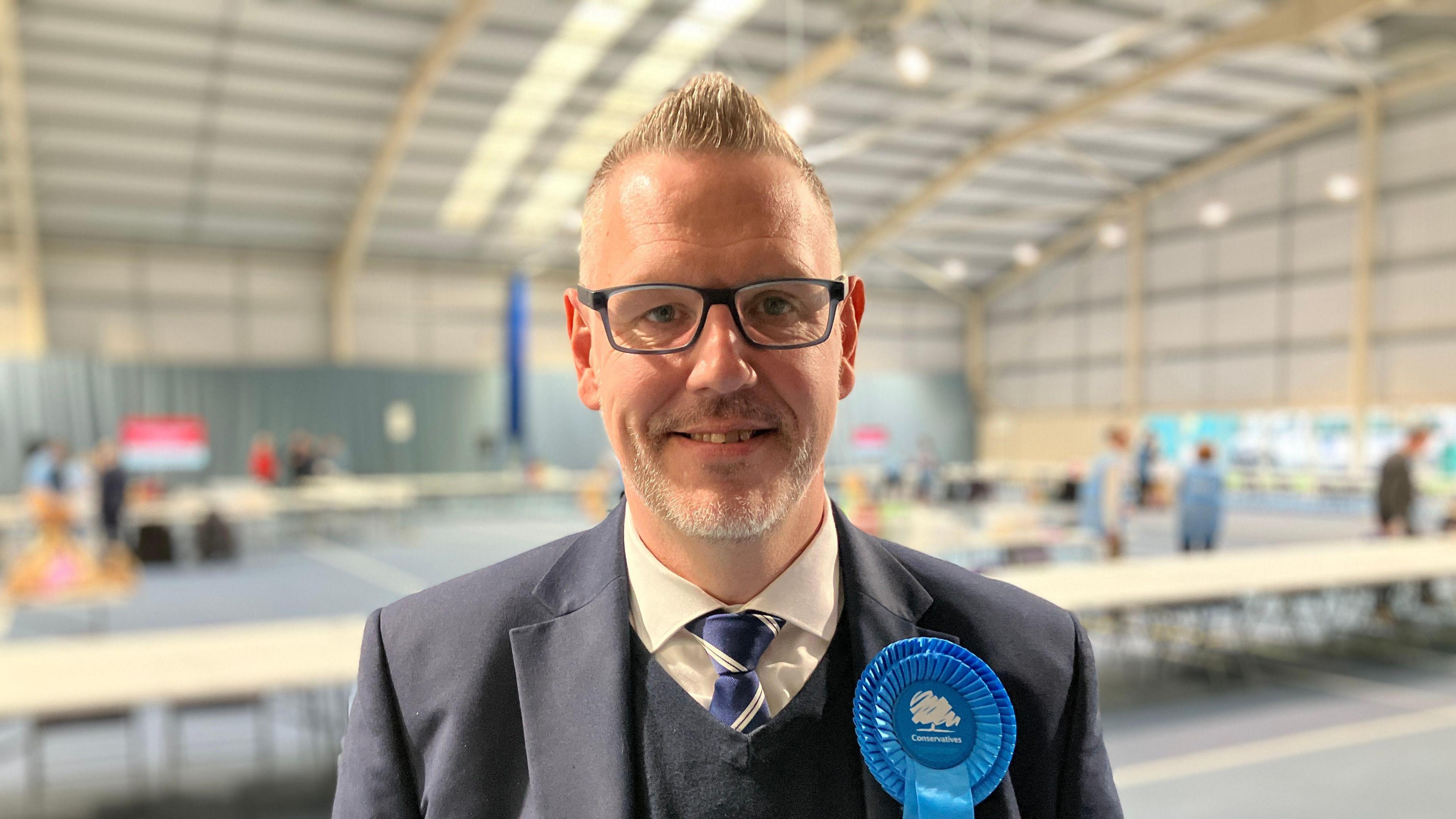 A man in a navy blue coloured suit and blue V-necked jumper with a blue tie with white diagonal stripes is pictured in a sports hall with courts and people behind him. He also has a blue Conservative rosette on his lapel and black glasses.