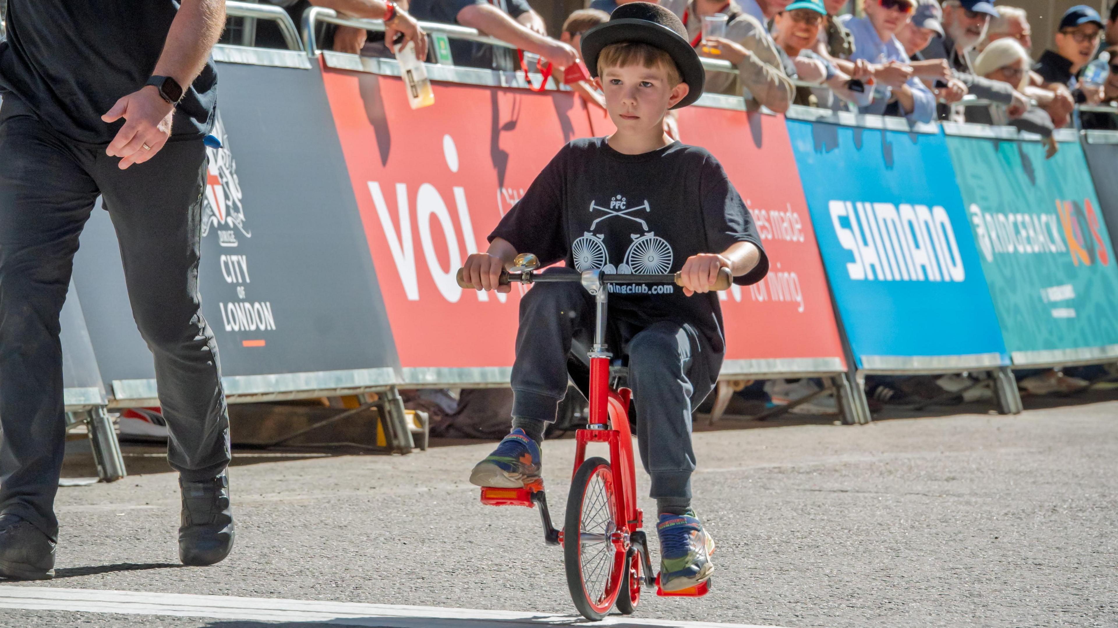 a boy in a bowler-style hat, jeans and t shirt rides a red small penny farthing bike on a sunny day as crowds look on behind him. he is joined by and adult, jeans just seen, by his side.