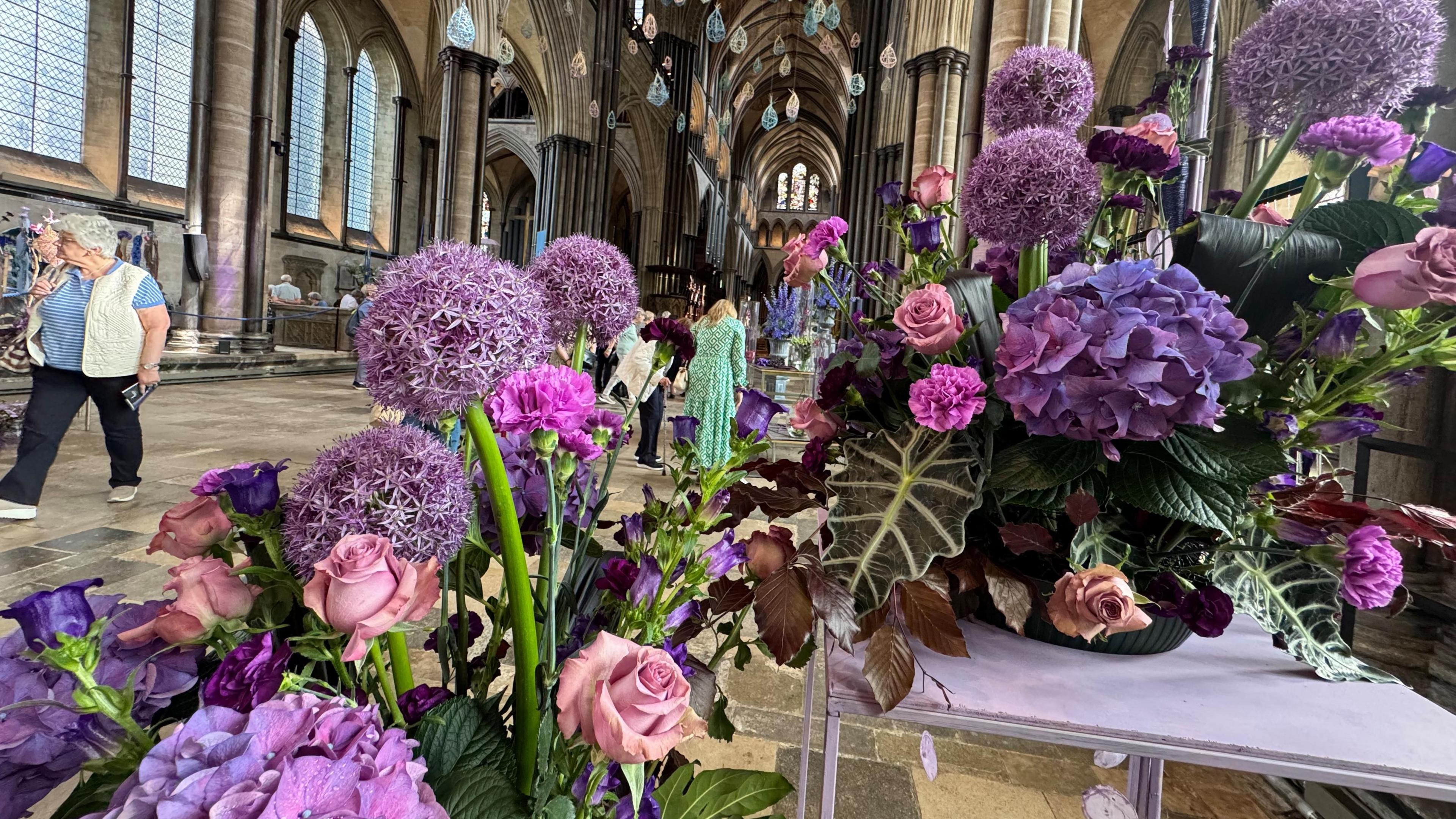 An arrangement of purple and pink flowers on two white tables. You can see people in the hall walking around looking at things in the background.