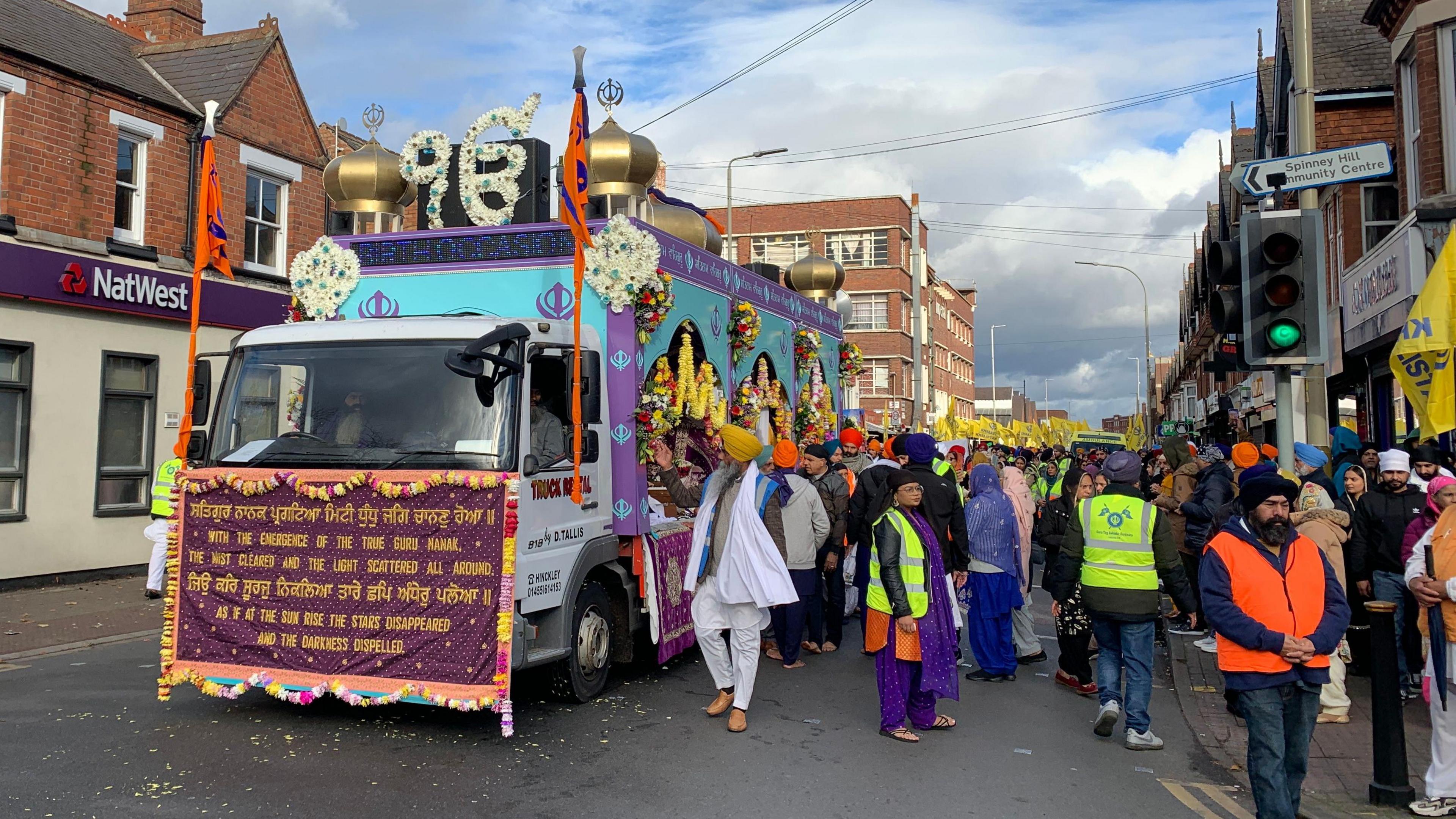 A lorry with a purple banner on the front with gold text. The main bulk of the lorry is turquoise and purple with colourful flower wreaths attached to the side and gold domes on top. Crowds can be seen behind the lorry on the road.