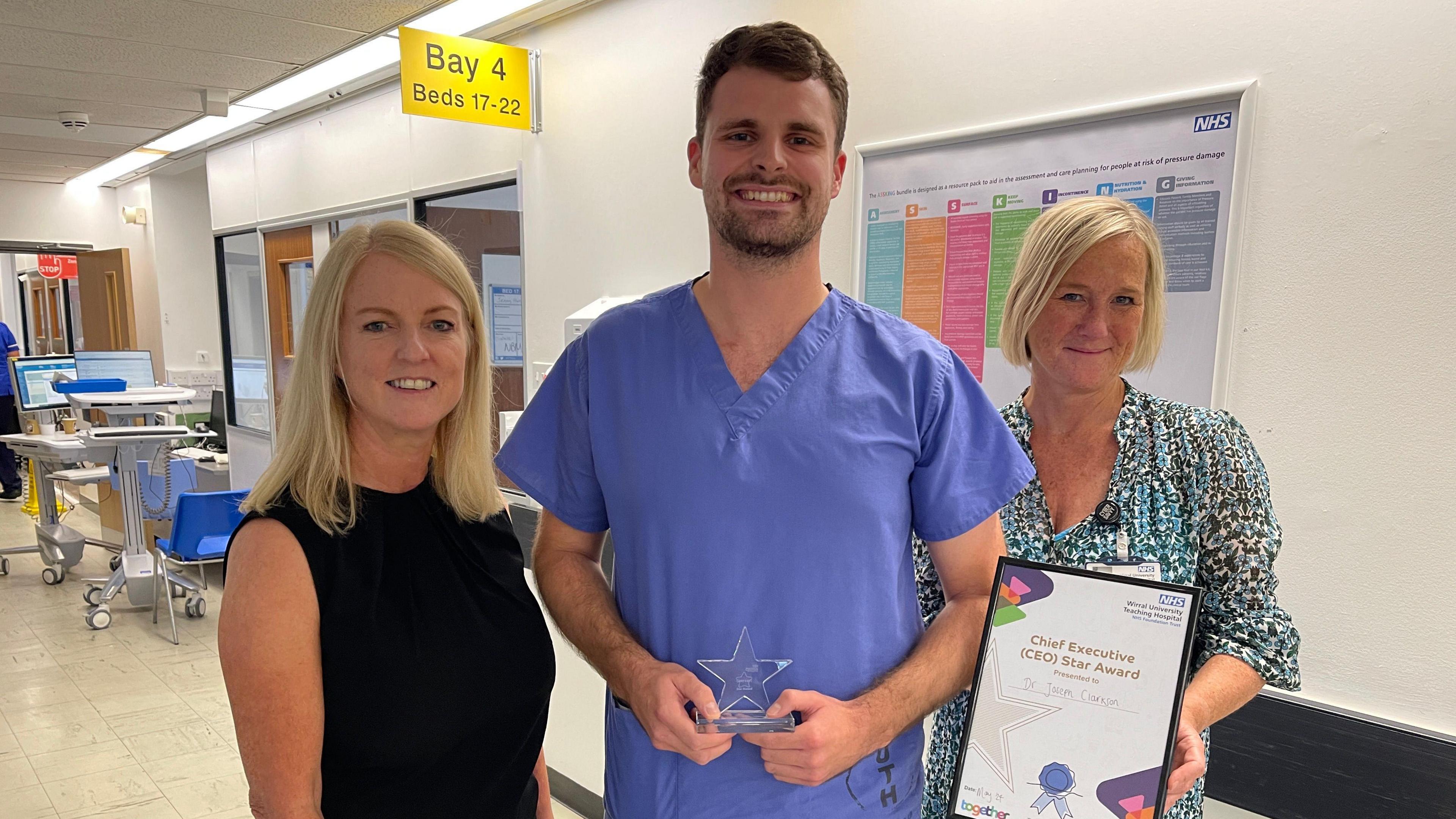 Joe with Dr Nikki Stevenson, Executive Medical Director (left) and Janelle Holmes, Chief Executive at Wirral University Teaching Hospital (right).
Joe is wearing blue doctor overalls, holding the star trophy, as the trio stand in a hospital corridor