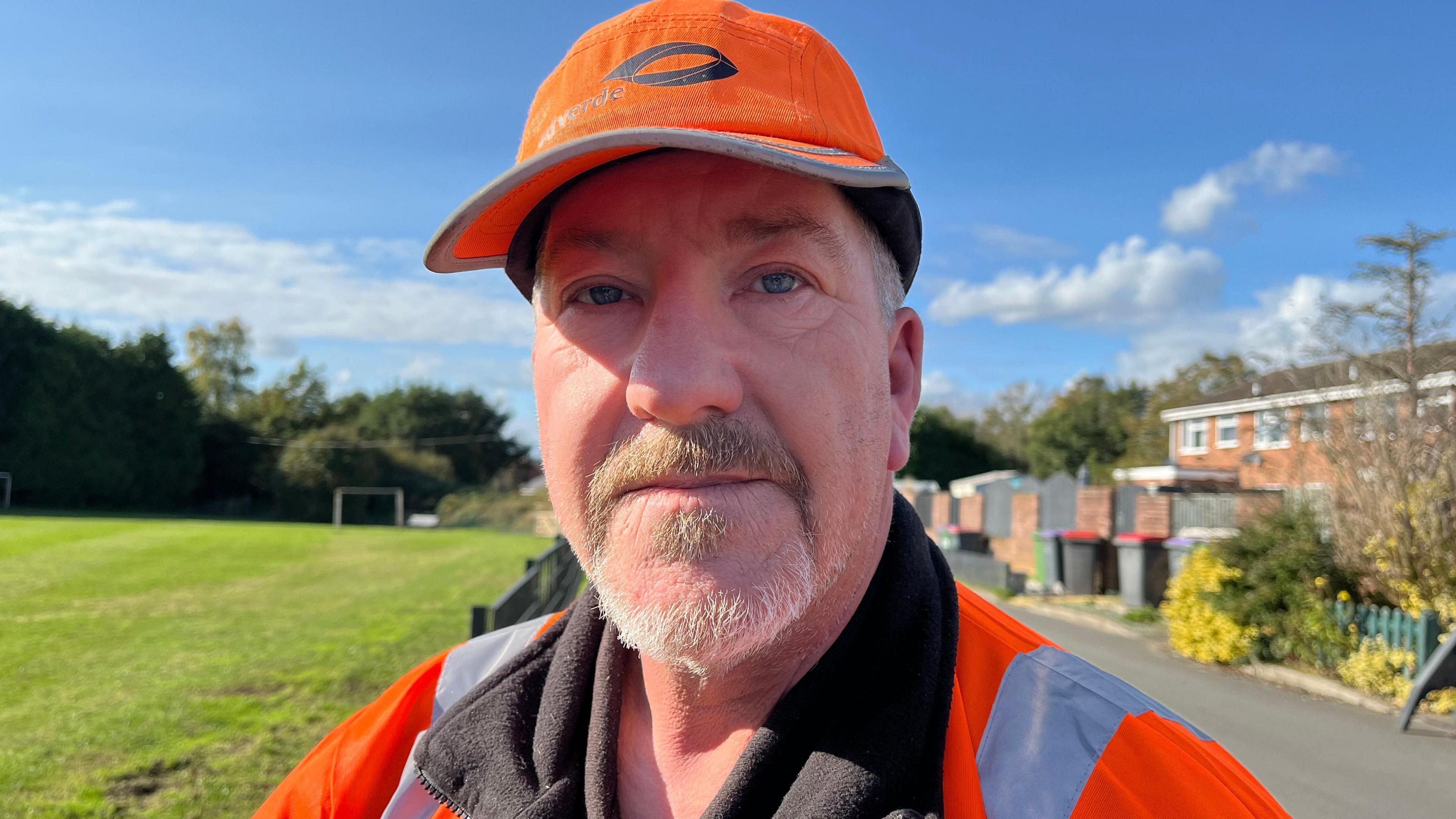 A man with short greying hair, moustache and stubble. He is wearing hi-vis jacket and branded baseball cap and standing between a playing field and row of houses.