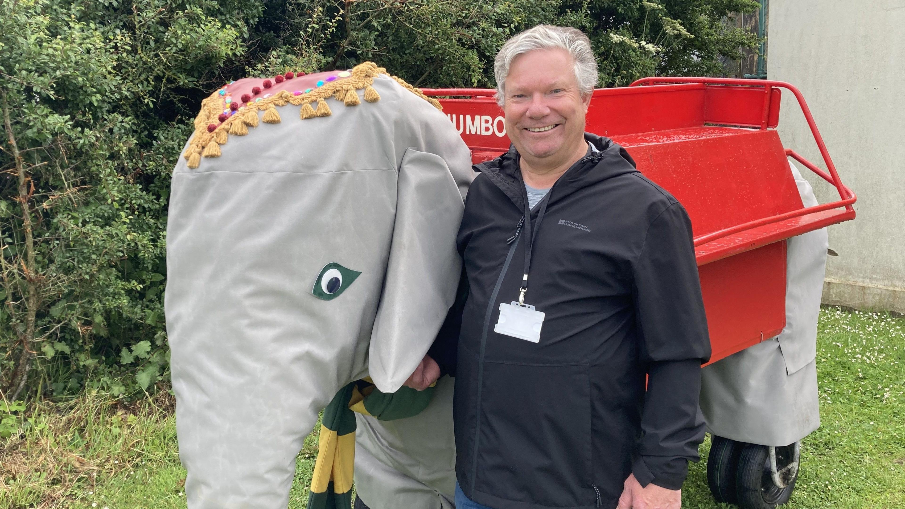 A man with grey hair and a black waterproof jacket standing next to a grey mechanical elephant which has a red seat on the top. 