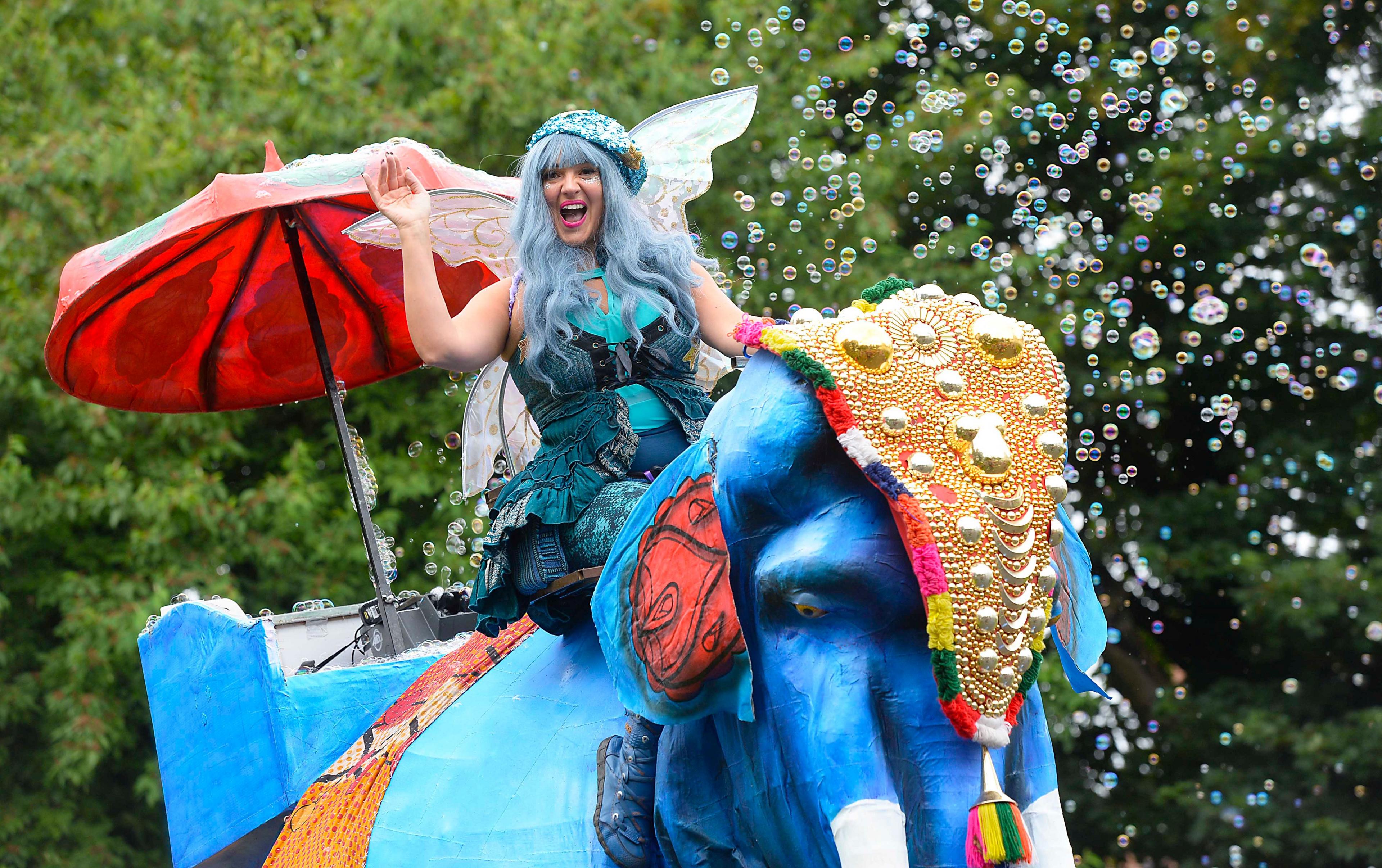 A woman, dressed in blue with blue hair and fairy wings, riding on a large blue elephant puppet, blowing bubbles into the air