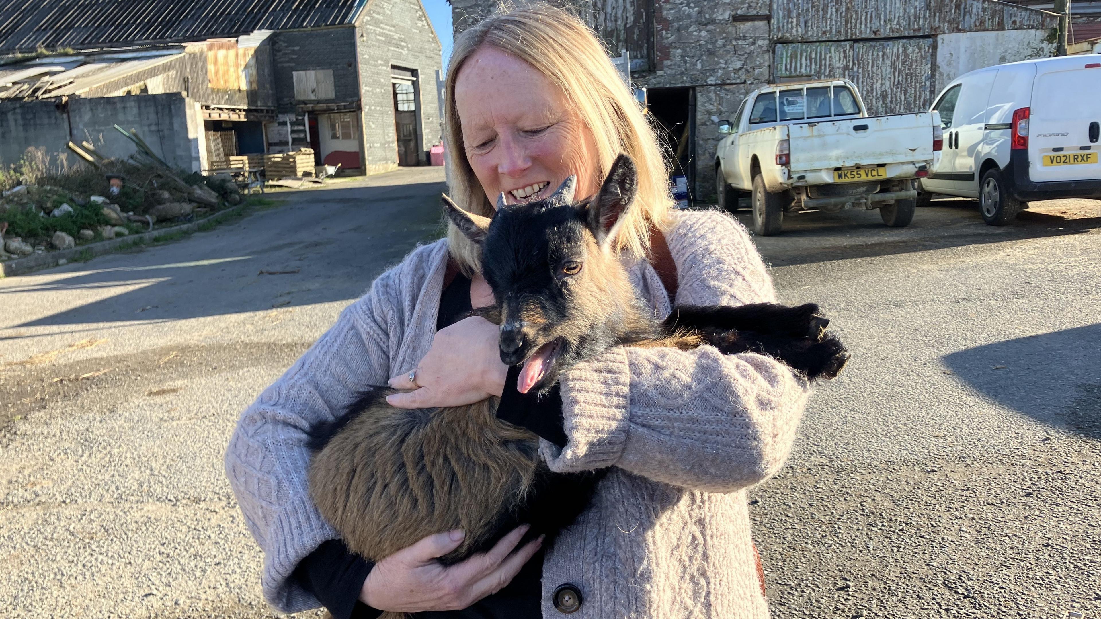 A woman in a grey jacket hugs a pygmy goat who has its tongue out. There are cars and farm buildings in the background.