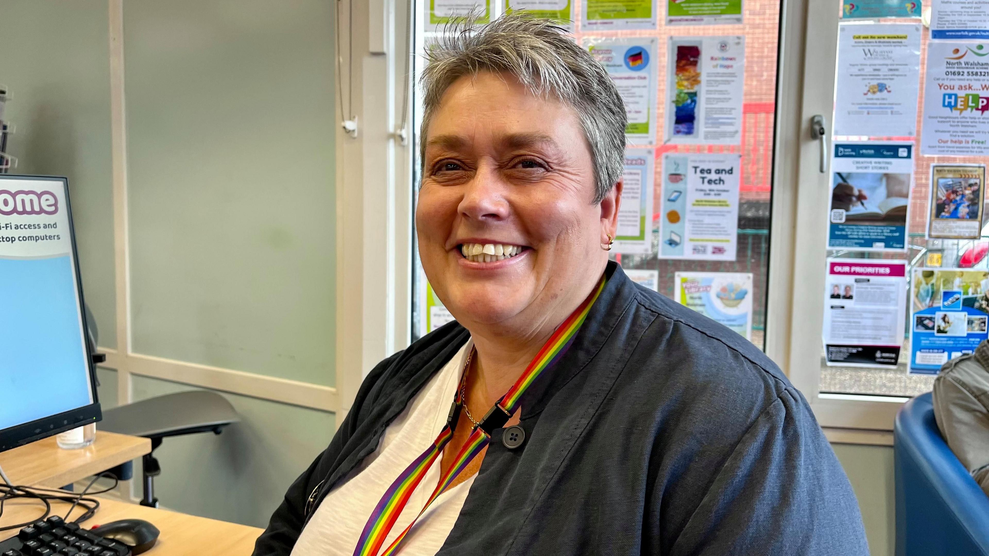 Gail Harvey is sitting at a computer and smiling at the camera. She has short grey hair and is wearing a white T shirt and a blue jacket. She has a rainbow lanyard around her neck.