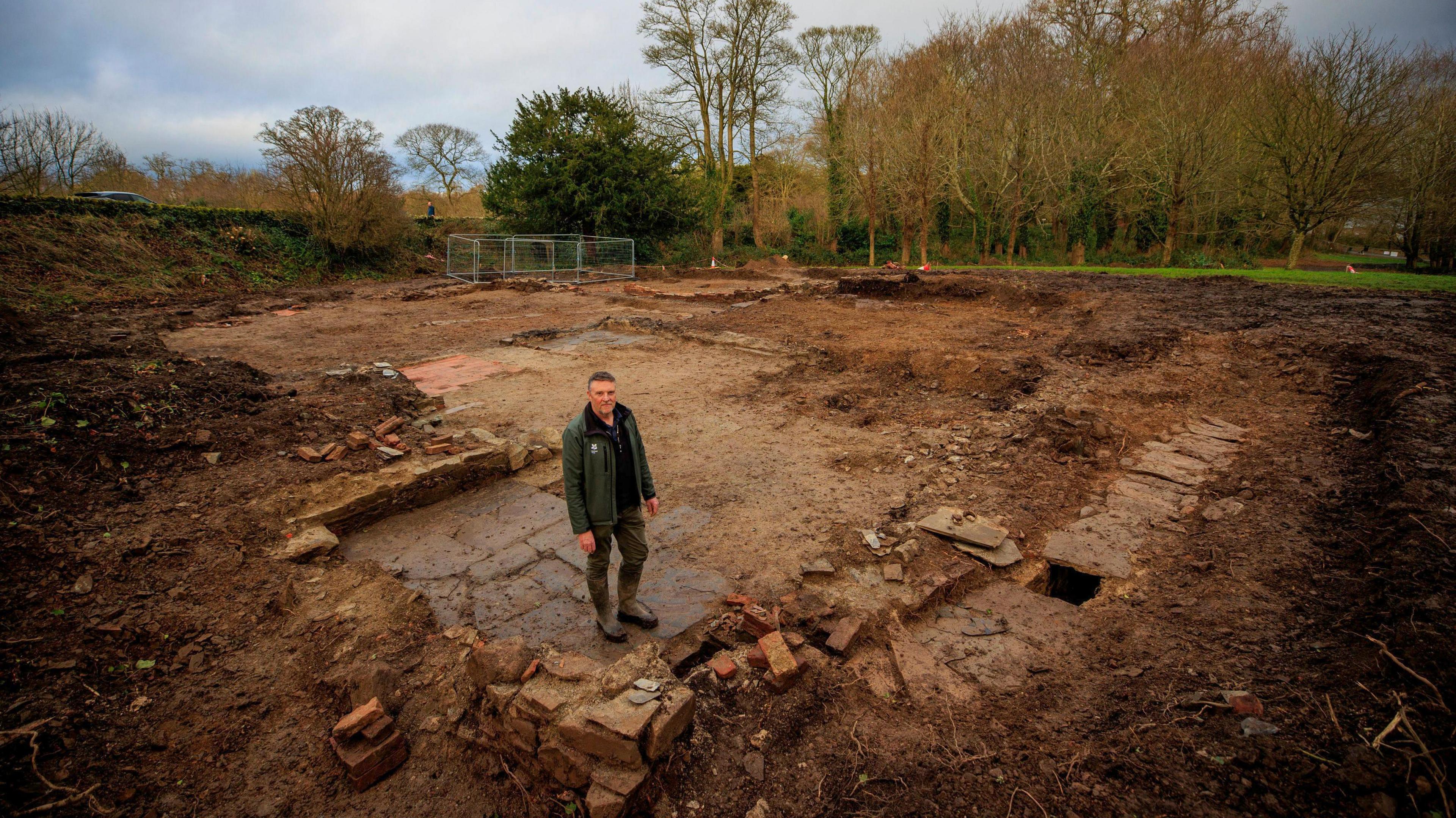 Malachy Conway wears a green jacket, dark trousers and boots as he stands in the middle of the site which has been unearthed. Bricks, tiles and stones are visible which were once covered in dirt.