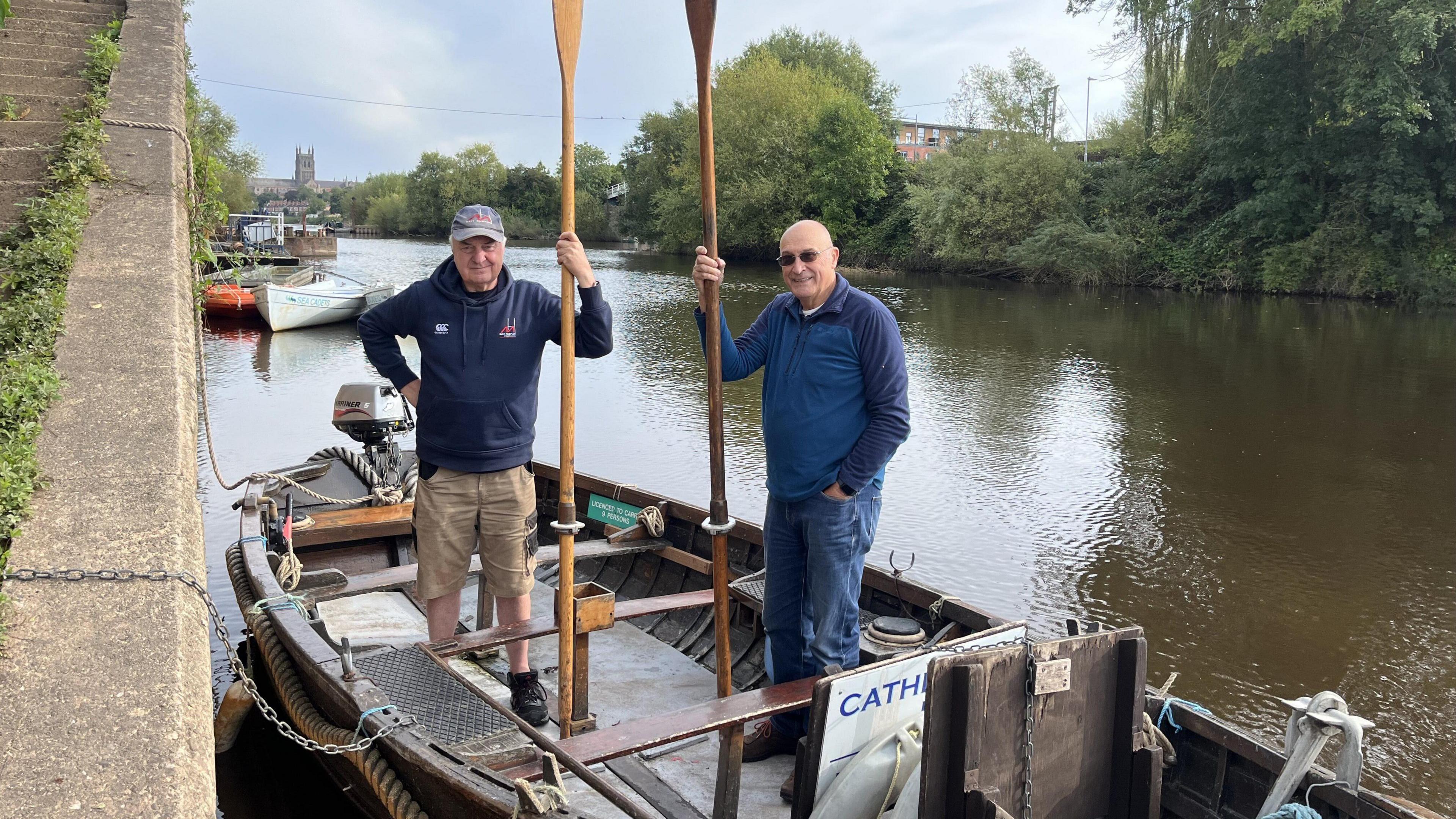 Two men standing on the wooden ferry on the river holding an oar each.