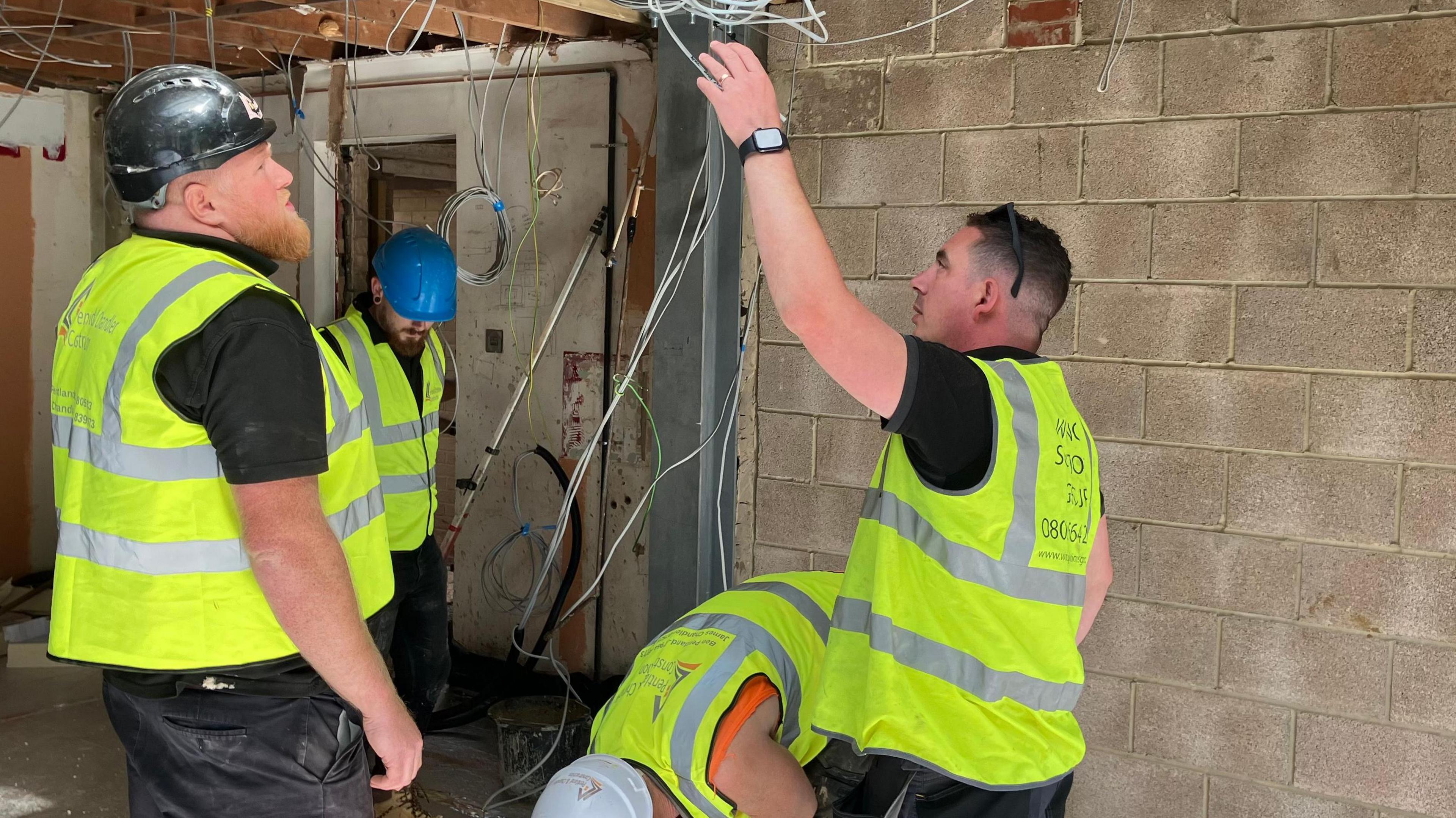 Three workmen in high visibility jackets inspect wiring on a building site. 