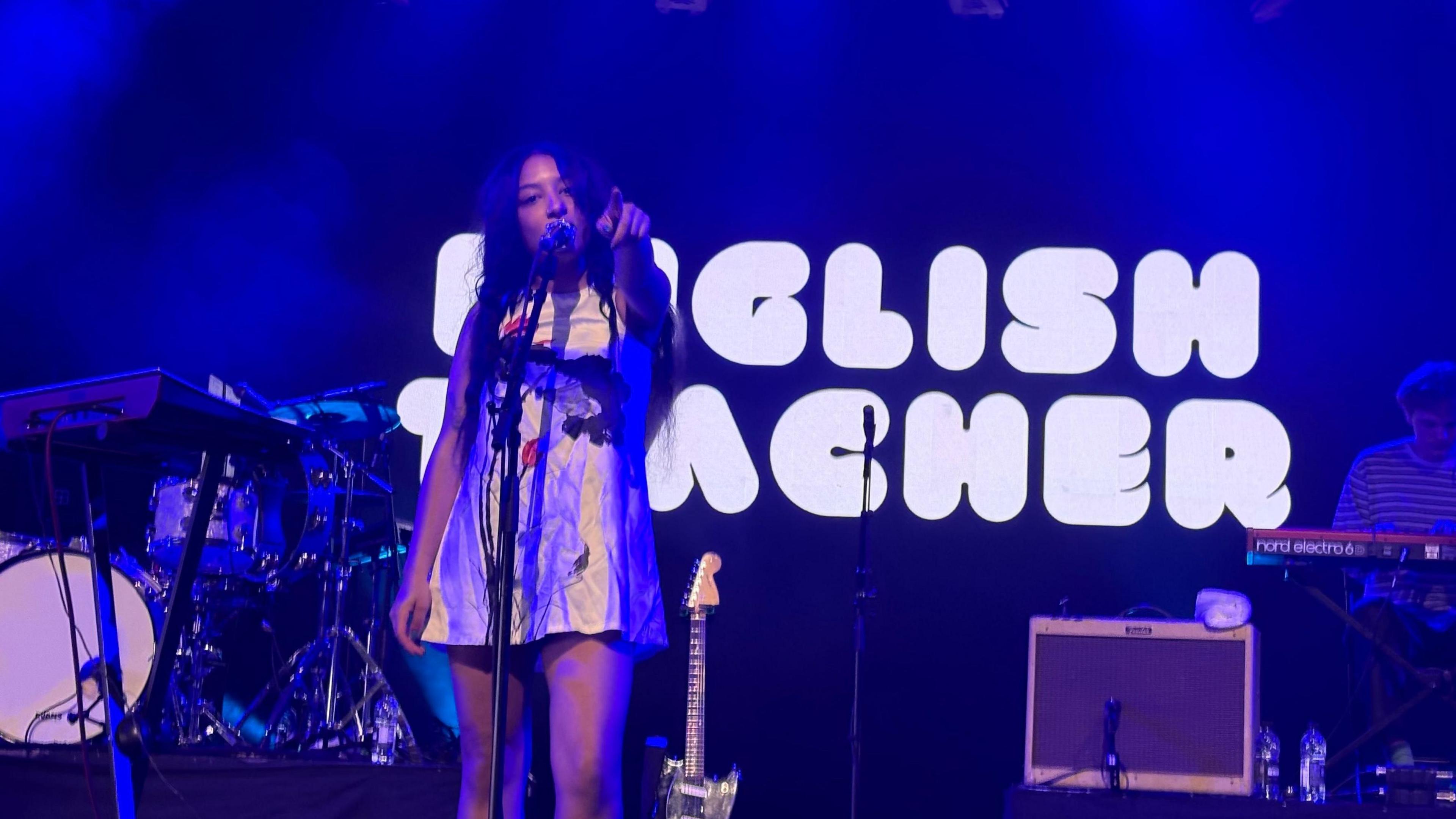 The lead singer of English Teacher has long dark hair and is wearing a short silver dress. She is singing and pointing to the crowd. The band's name is emblazoned across the back of the Big Top stage.