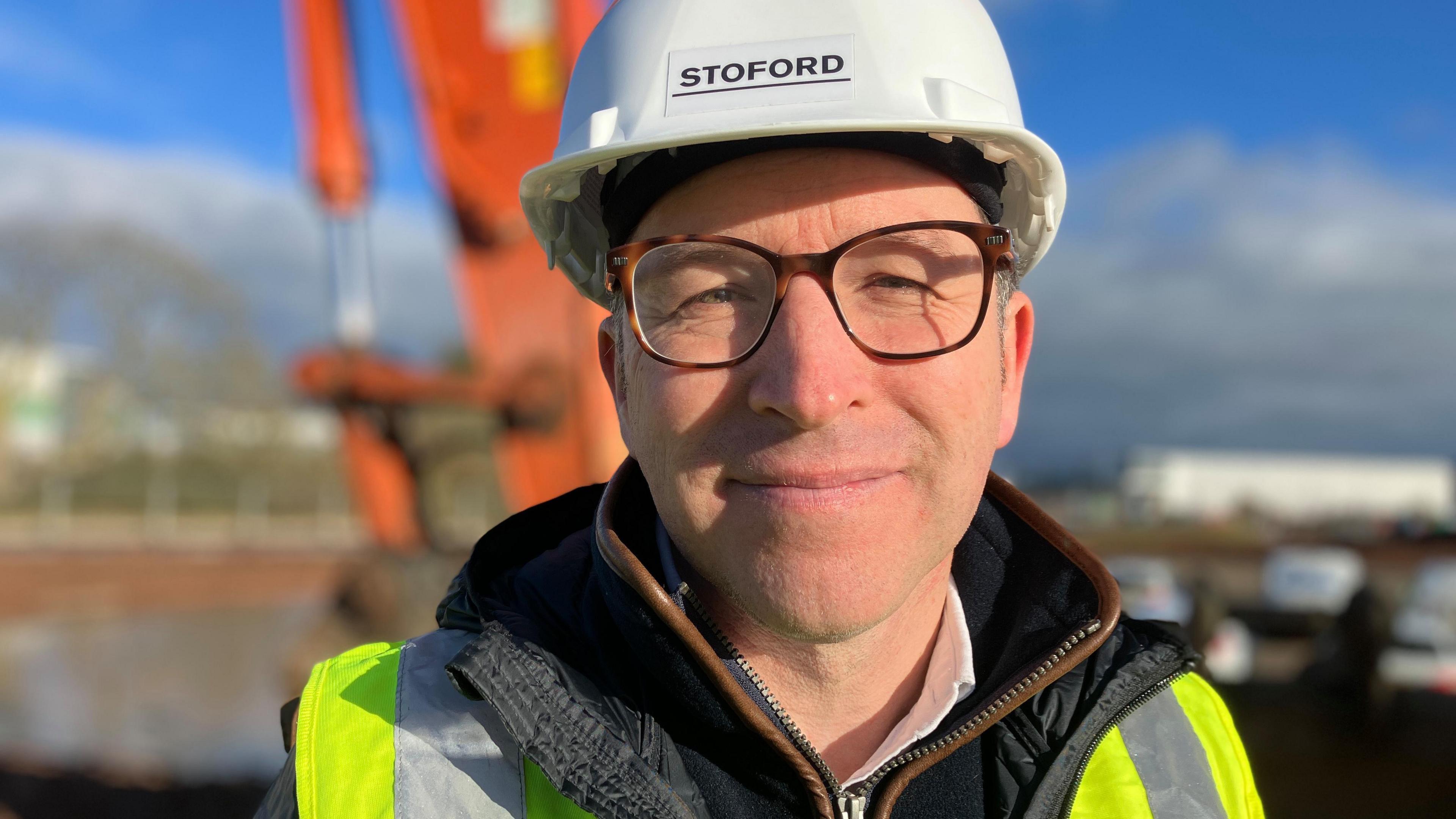 A man wearing glasses, a hi-vis jacket and a white helmet emblazoned with "STOFORD" smiles at the camera. An out-of-focus building site is visible behind him.