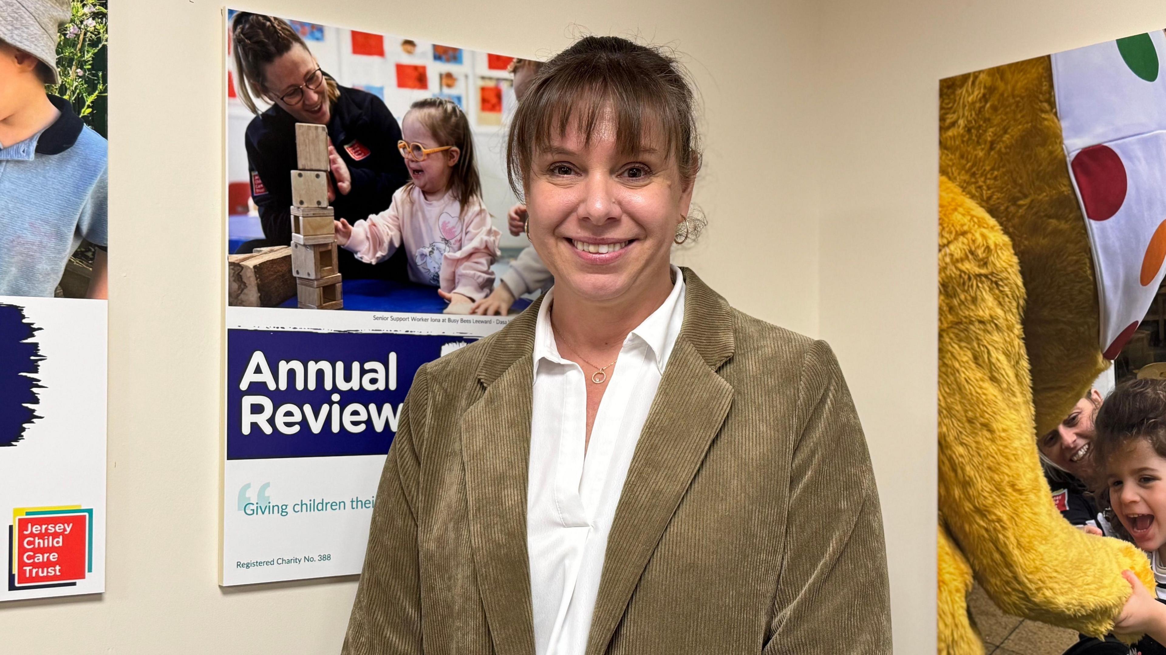 A woman in her late 30s with brown hair which is tied up. She has a block fringe and wears a white shirt and green corduroy jacket. She is stood infron of posters promoting the work the JCCT does.