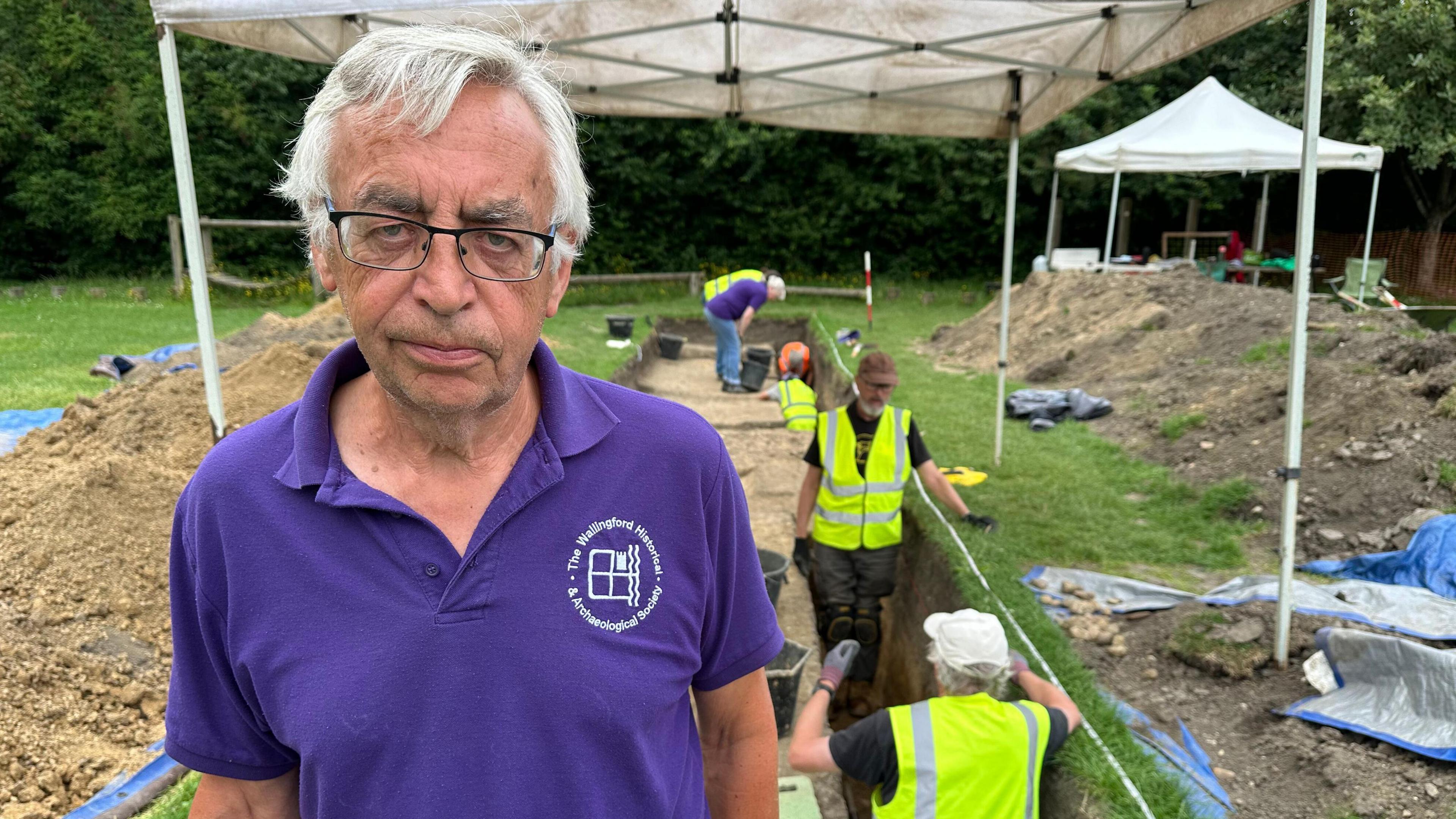 Steve Capel-Davies, stood in front of the digging site, wearing a purple shirt with the historical group's insignia