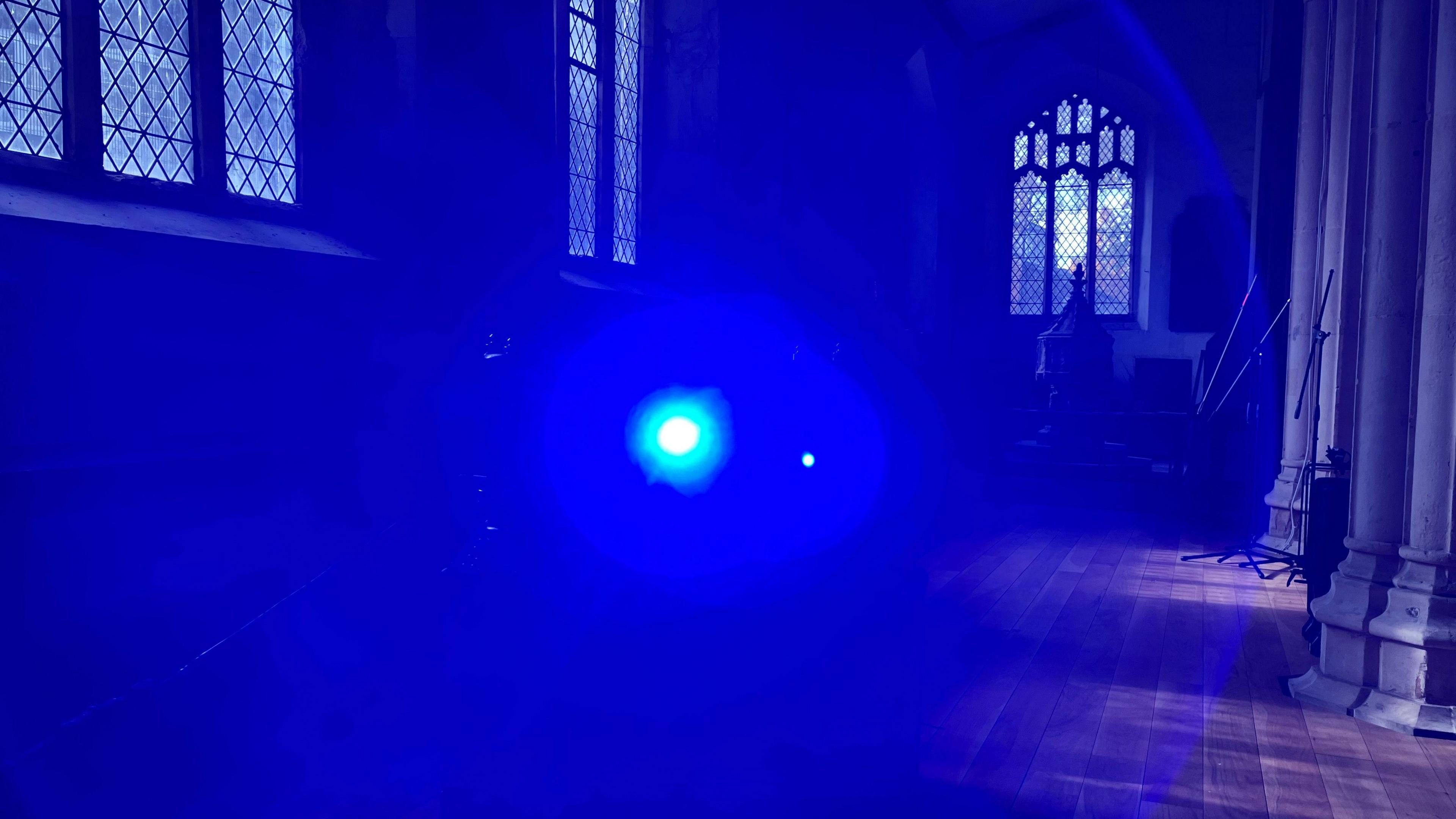 The blue light of a projector lens, with a circular glow around it. There are church columns and a stained glass window in the background.