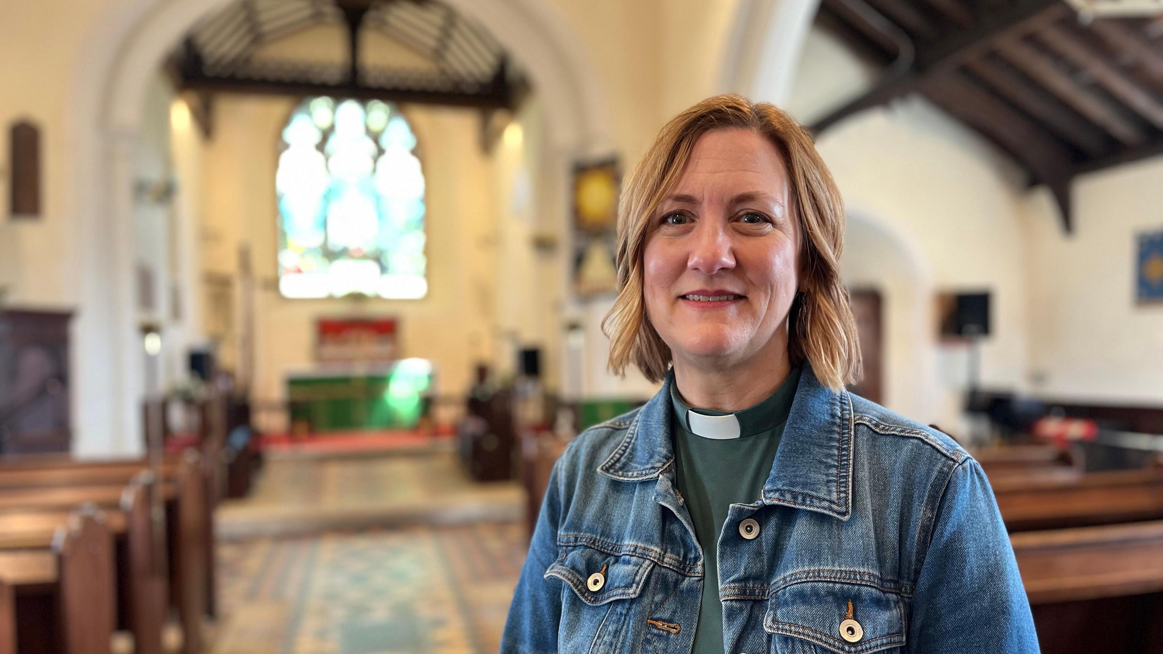 Reverend Elise Peterson inside St Mary and St Peter's Church in Wennington. The vicar has short blonde hair and is wearing a green top with a clerical collar and a denim jacket. In the background are rows of pews and a stained glass window.
