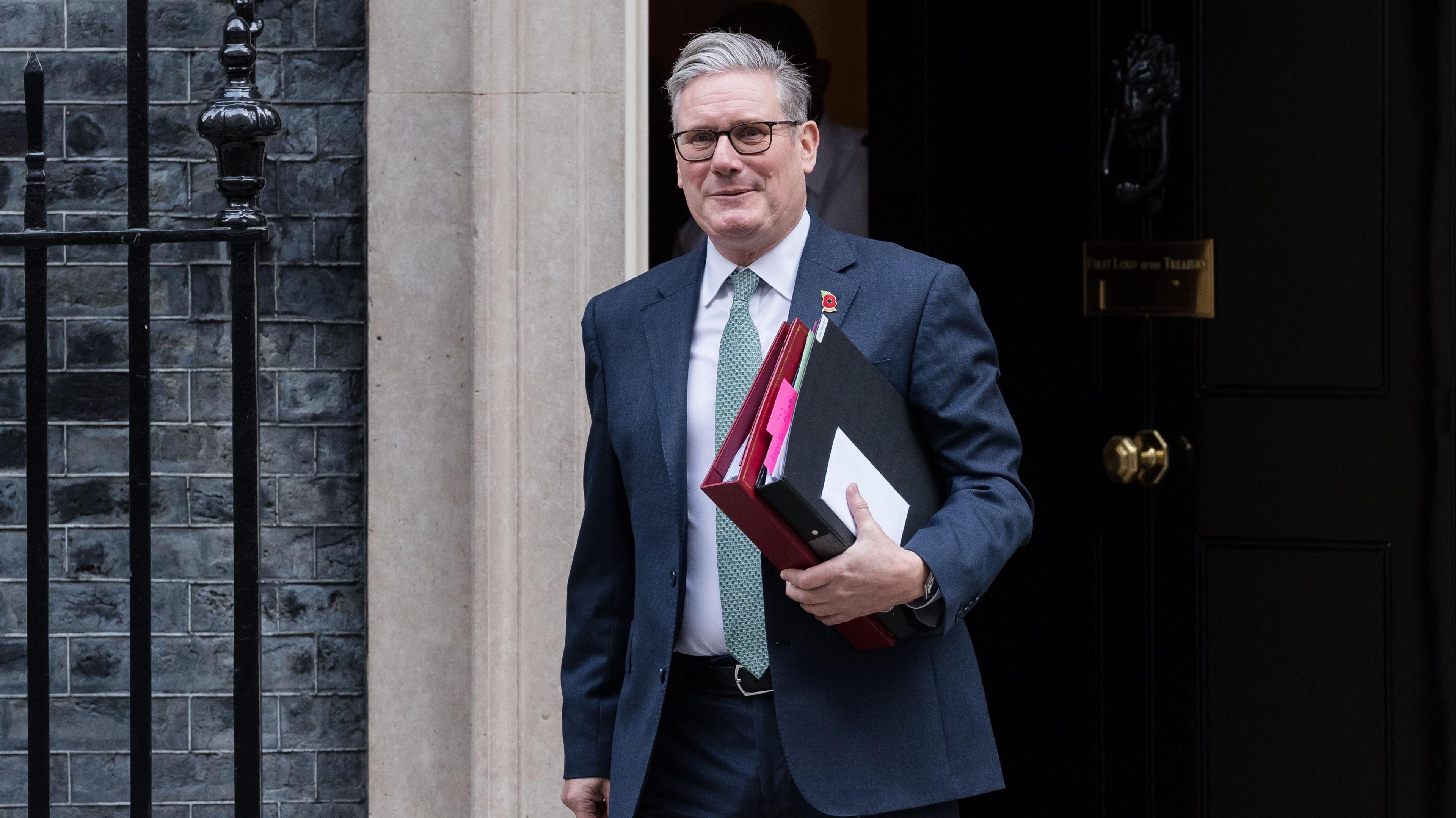 Sir Keir Starmer, wearing a dark suit, white shirt and light blue tie and carrying files under his left arm, walks out of 10 Downing Street.