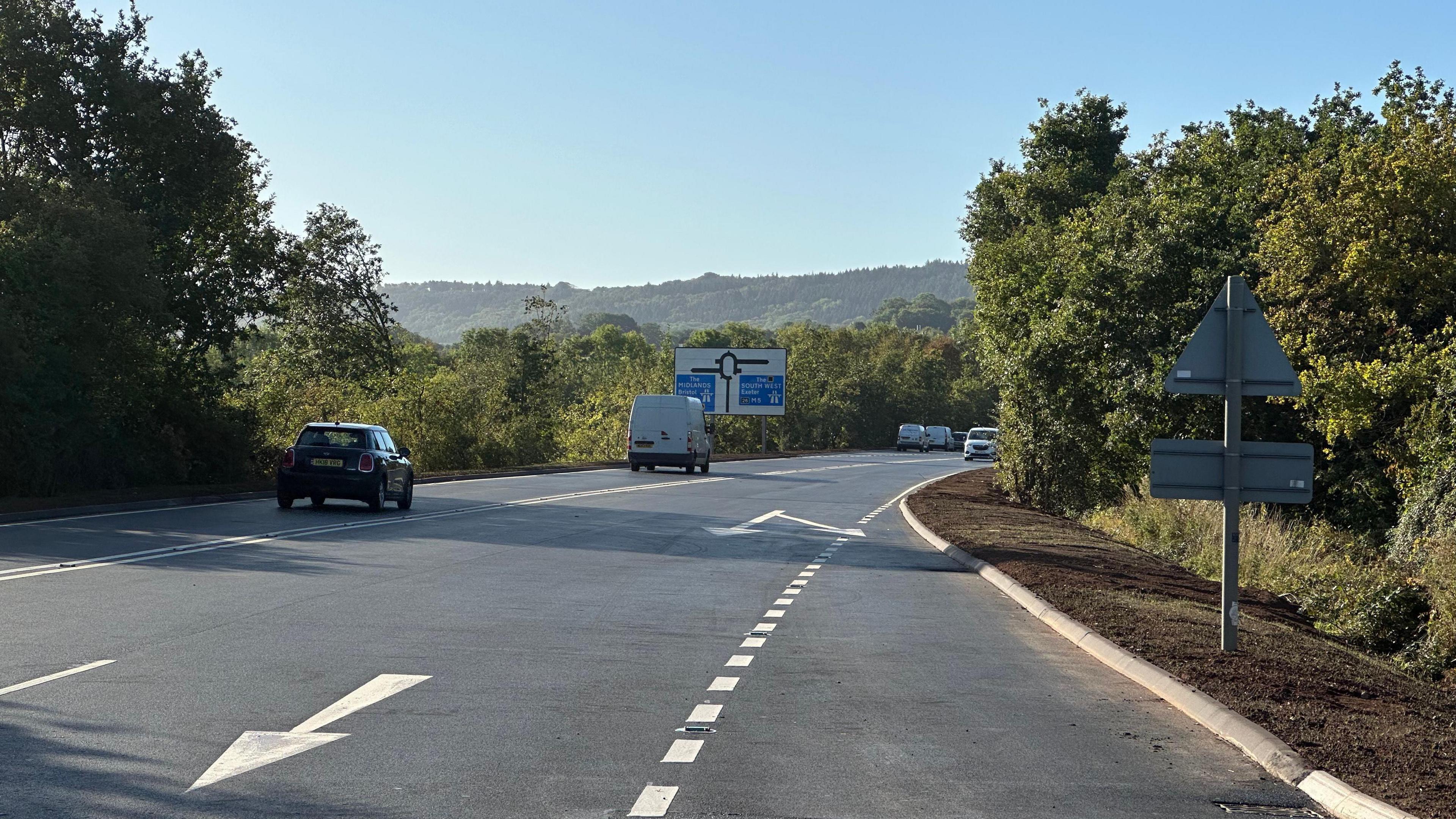 A road in Somerset. Several cars are travelling on the road. There is a road sign for a roundabout in the distance. Trees can be seen either side of the road.