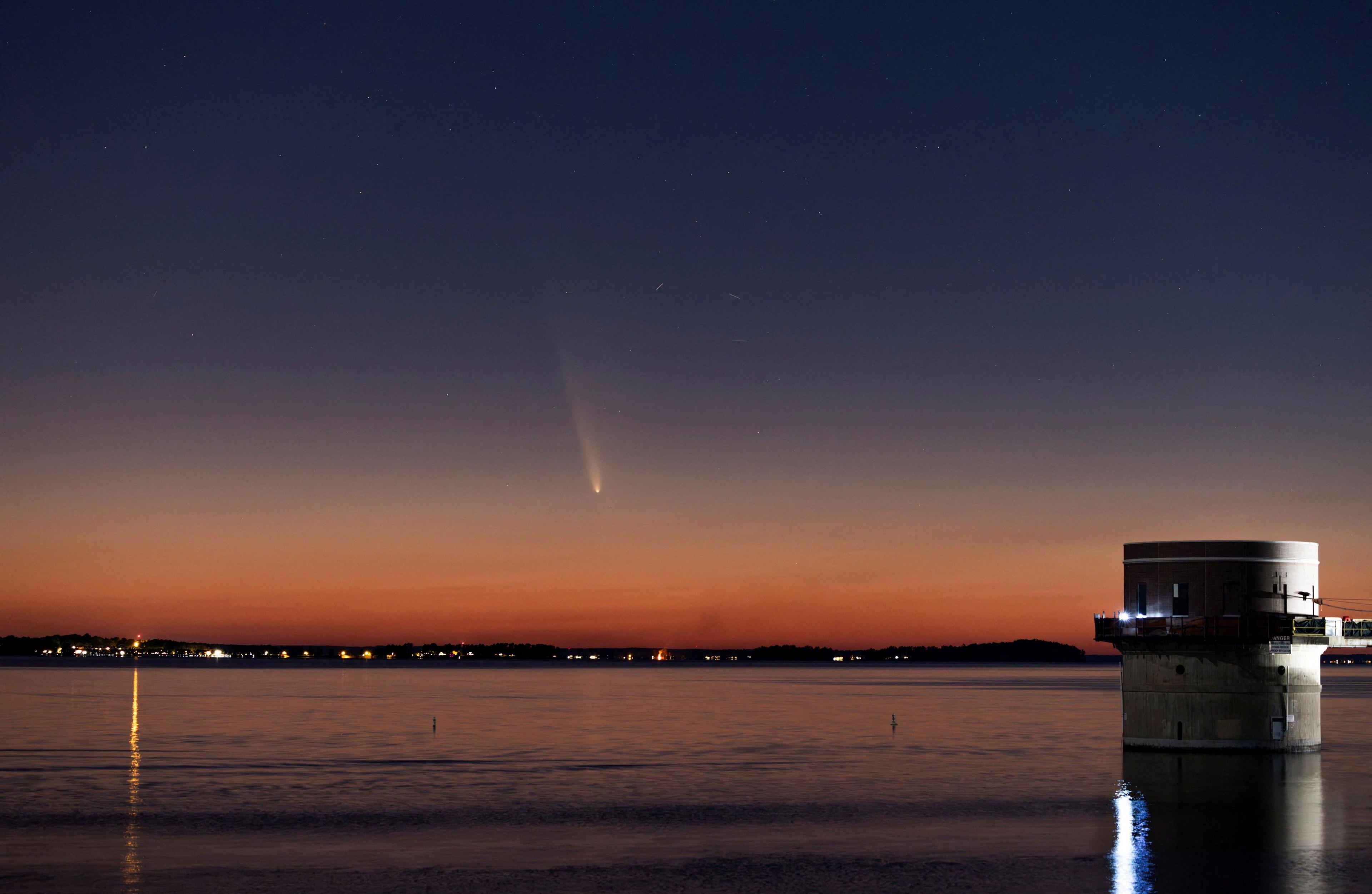 A building at the edge of a lake and then a settlement on the far side under an orange hue in the sky. Just above, in a bluer, higher portion of the sky, light from a comet can be seen.