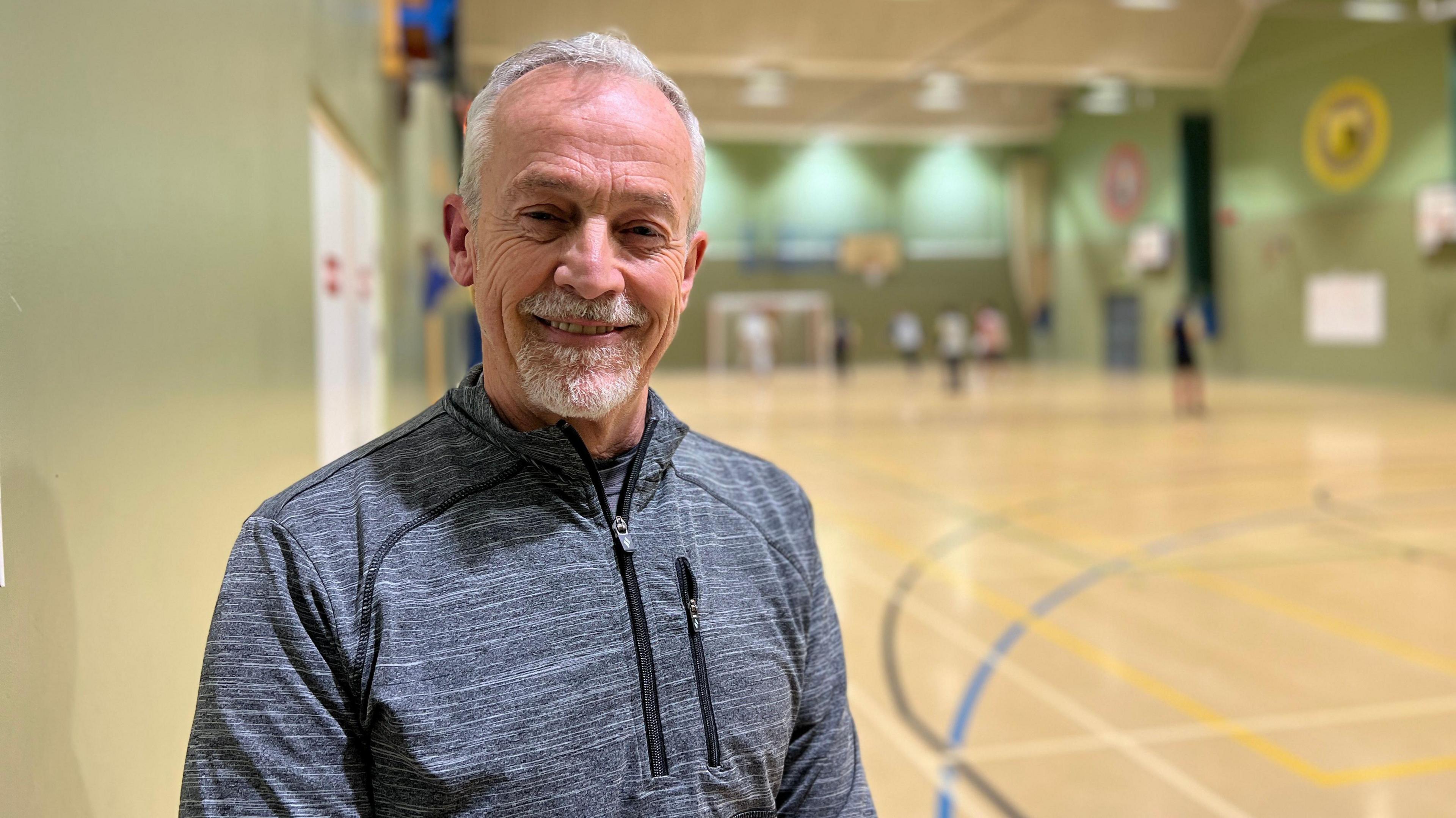 Gary smiles at the camera as the players are playing a game of futsal behind him. He is wearing a grey zip up fleece and has white, short hair and a white goatee.