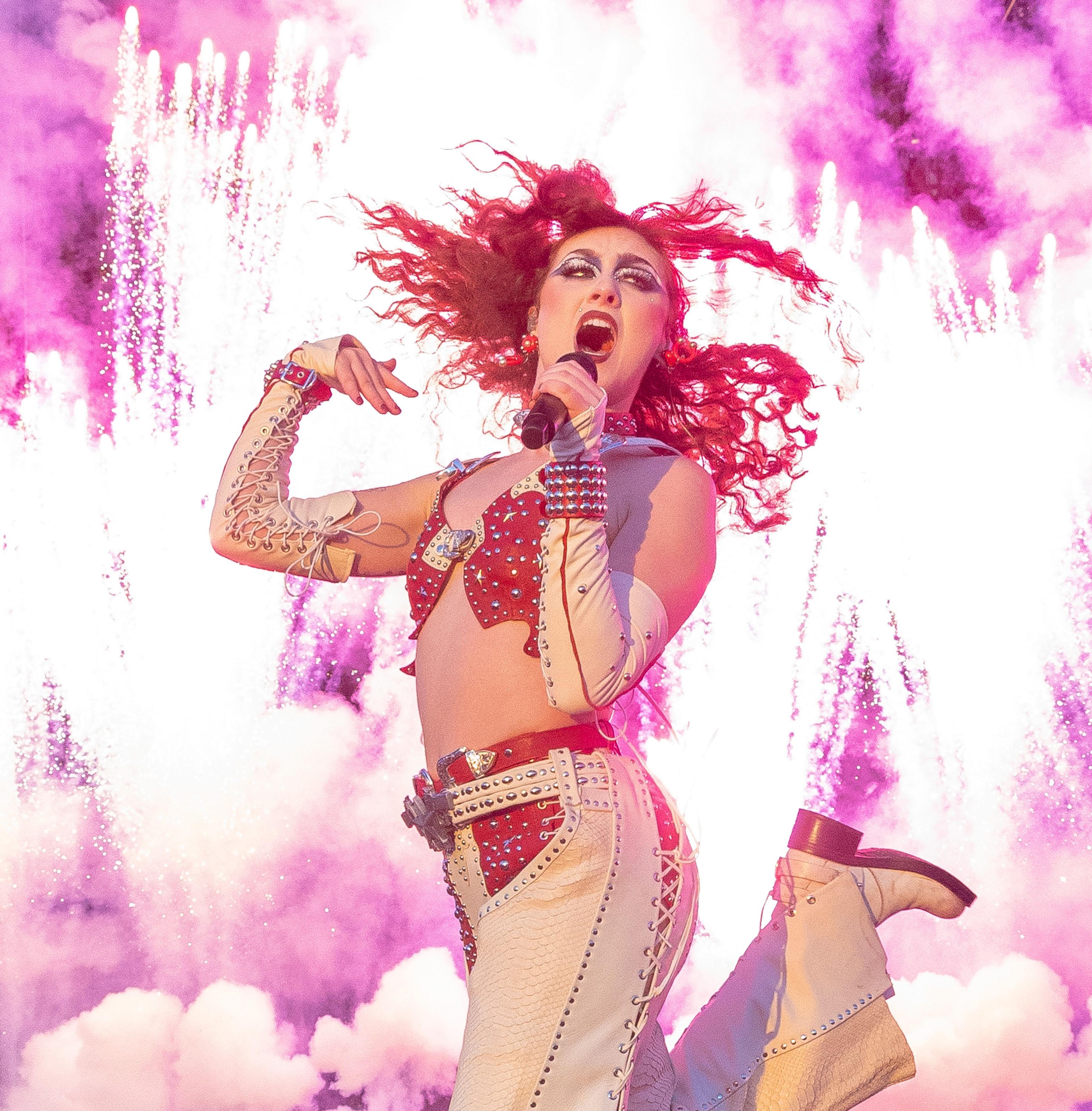 Chappell Roan in a white rhinestone cowgirl outfit kicks up her heels while pink fireworks explode behind her.