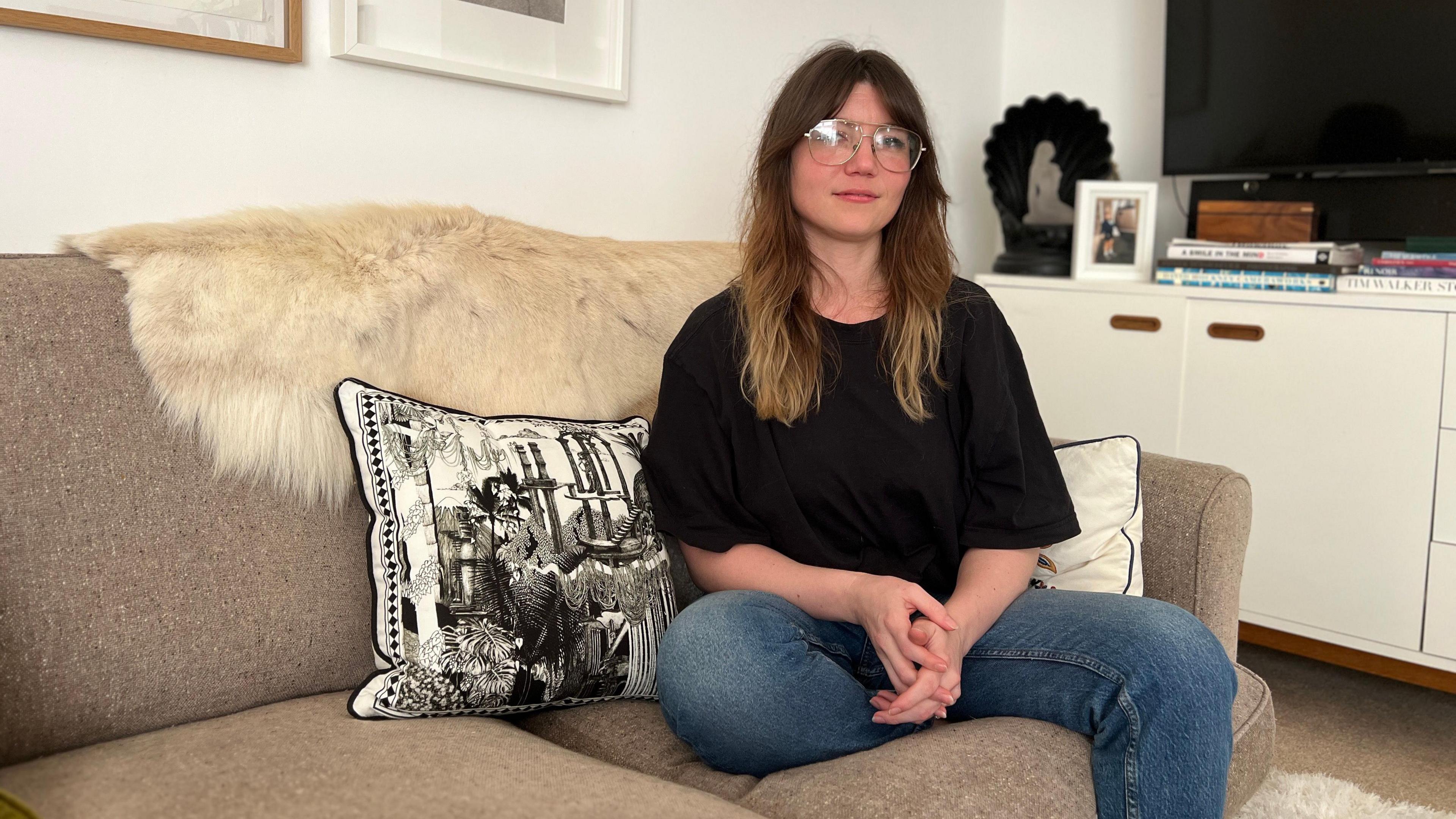 Abi looks at the camera as she sits on a grey sofa as she wears blue jeans and a black t-shirt. She has dark brown hair with lighter ends which comes just past her shoulders. She is sitting next to a cushion with black and white print pillow.