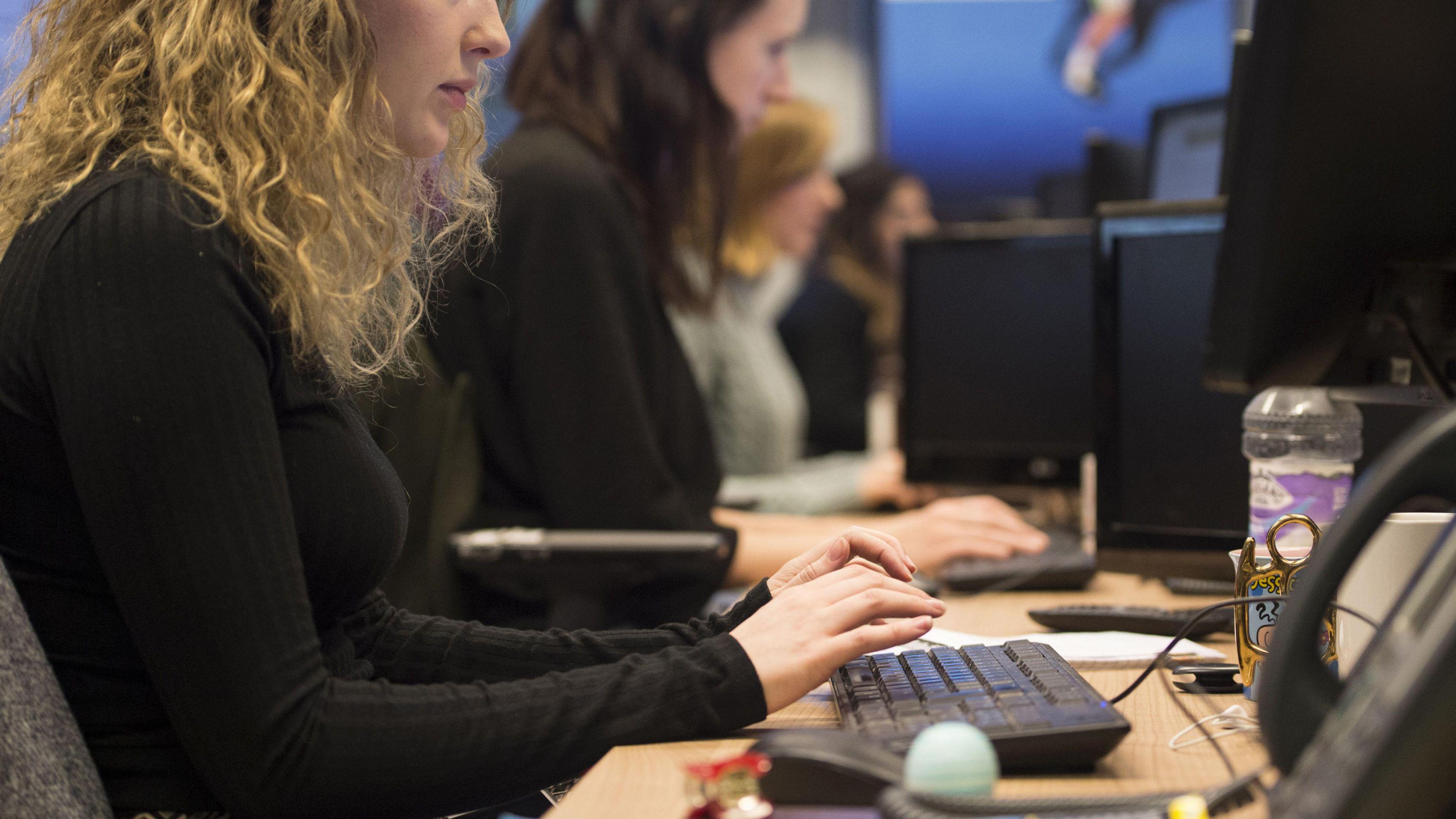 A woman working at her desk typing on a keyboard. A row of other workers blurred can be seen in the background.