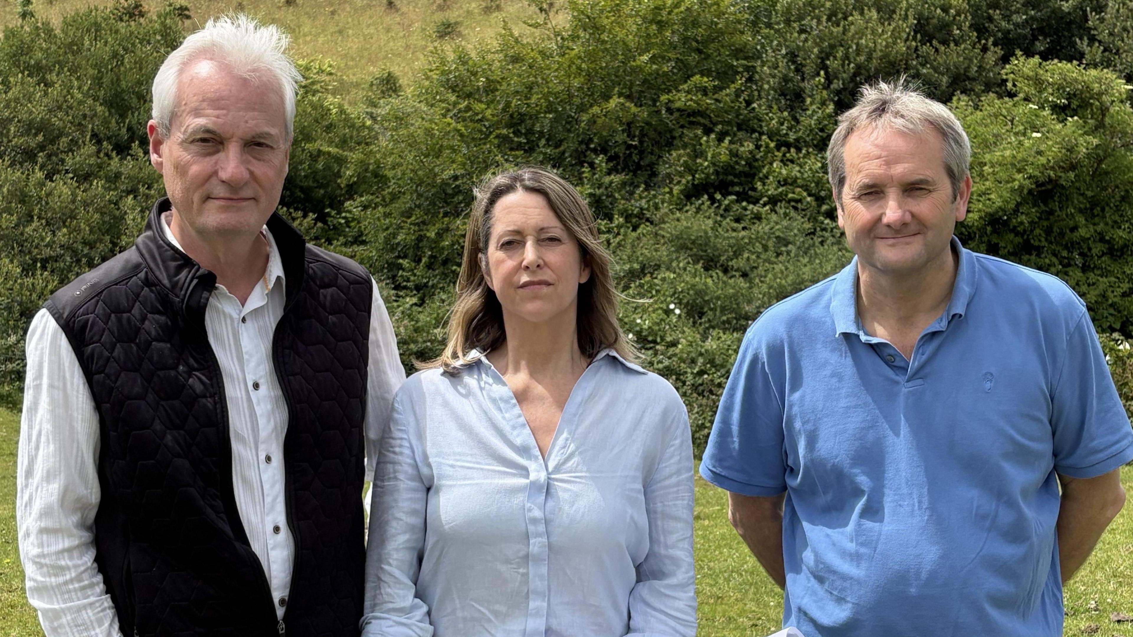 A woman with shoulder length brown hair wearing a blue shirt, standing between a man on her right with white hair, in a white striped shirt and black gilet, and a man with grey hair and a blue polo shirt on her left. They have some hedges and hills behind them.   