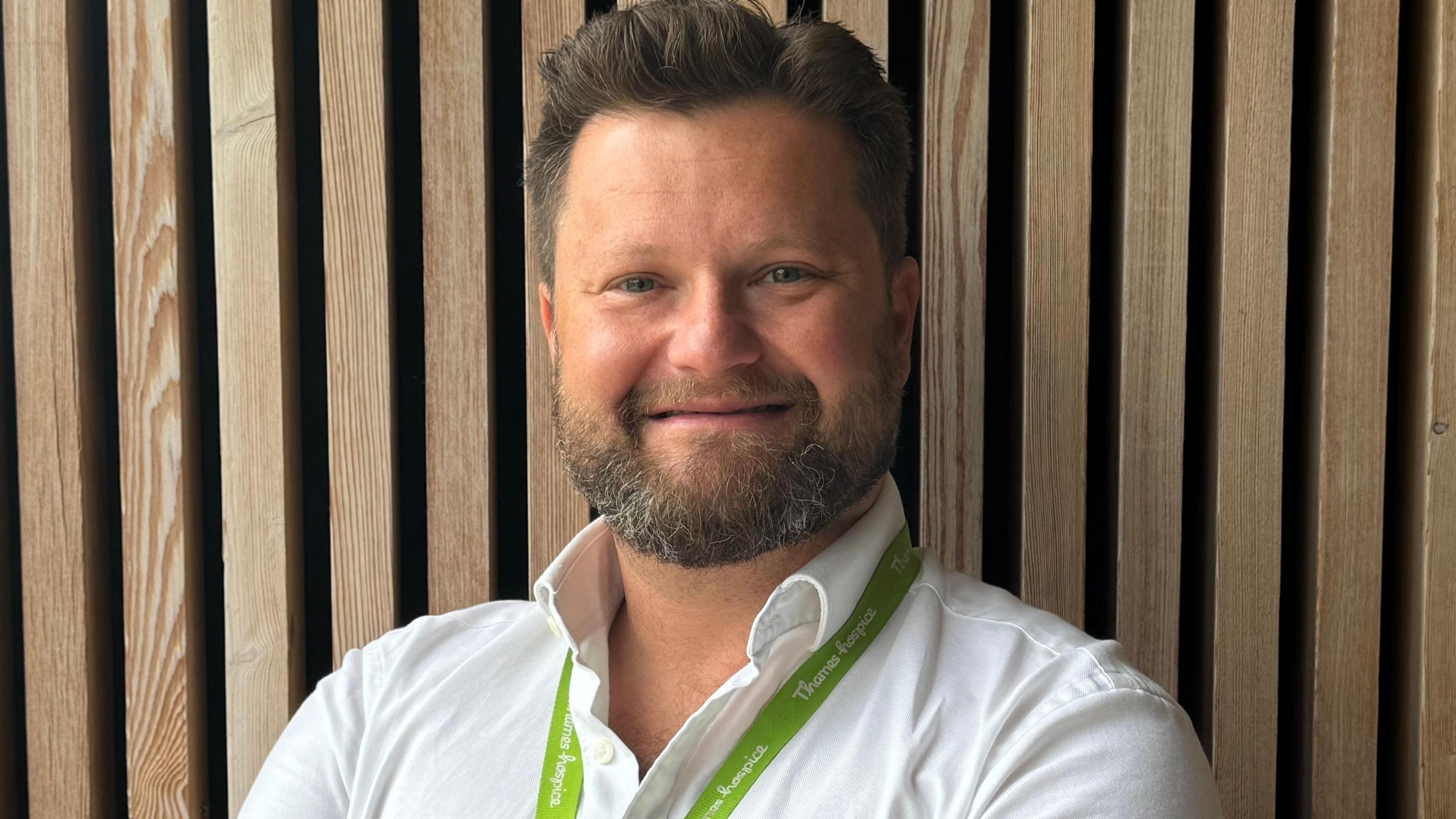 A man with brown hair and a beard flecked with grey, standing in front of a wooden wall and smiling, wearing a white shirt and a green lanyard.