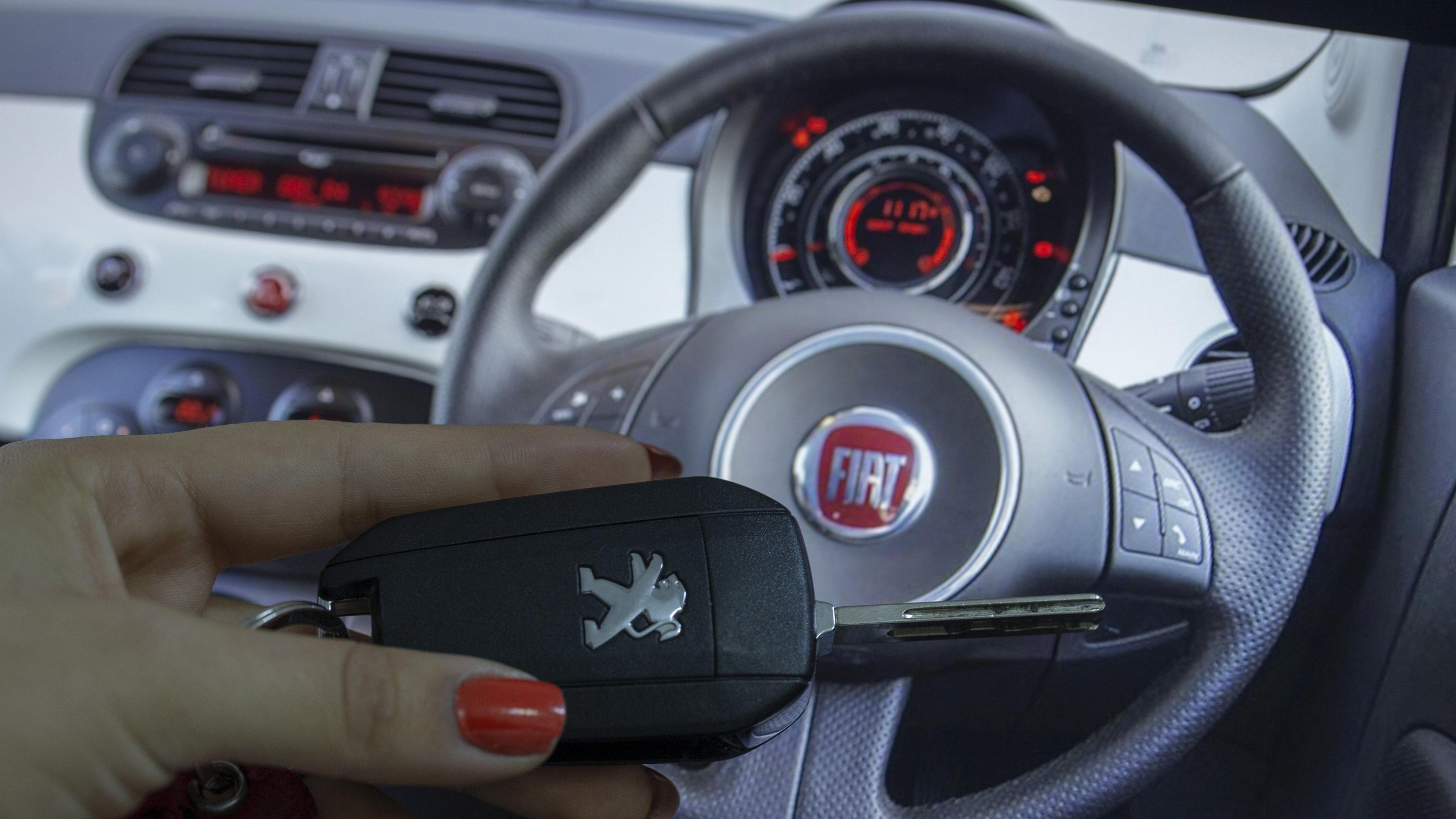 A hand with red painted fingernails holding a car key. The person is sat in a Fiat car and is holding the car key over the steering wheel of the car.